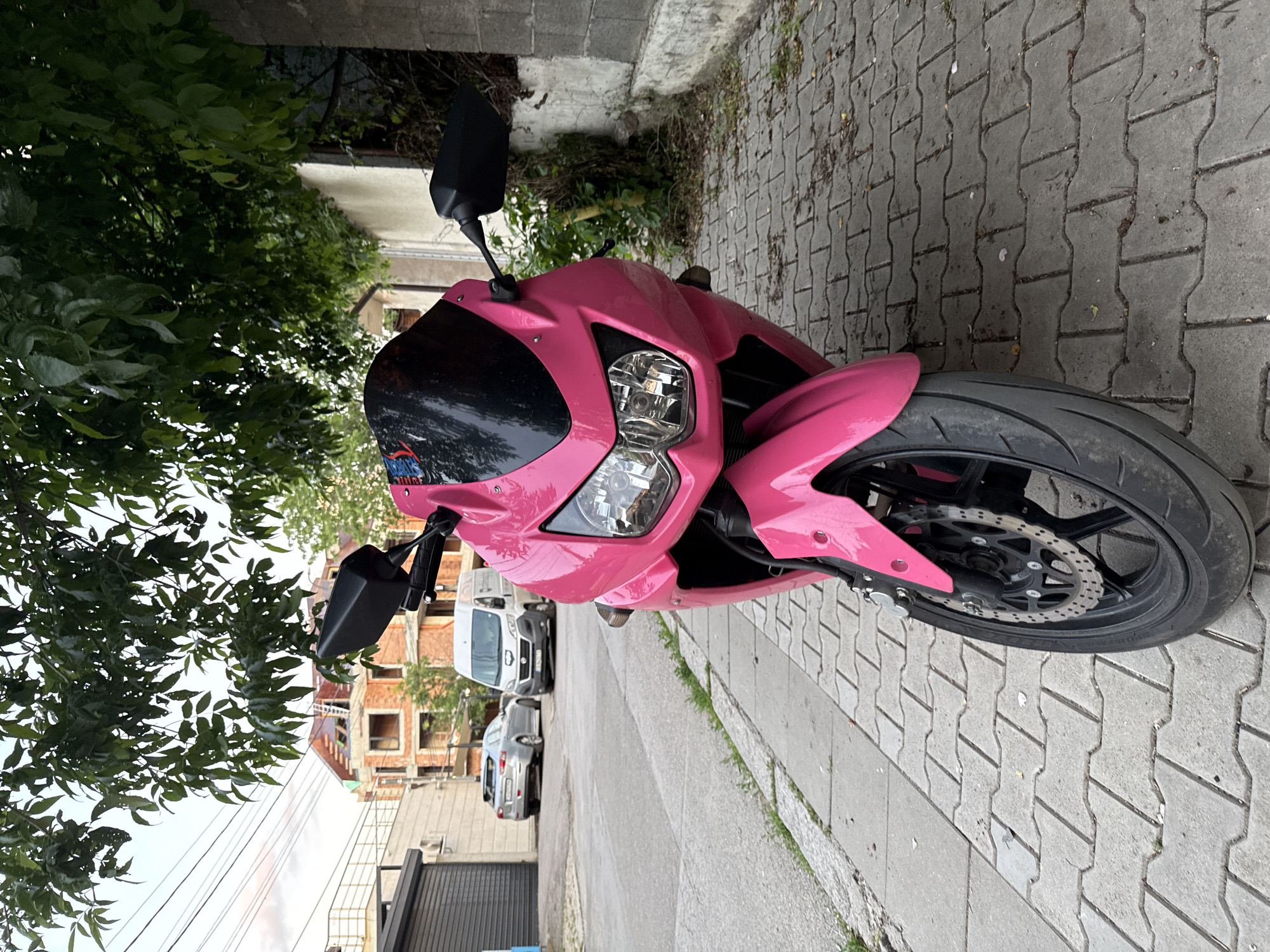 A pink sportbike is parked on a gray, interlocking paver sidewalk next to a tree with green leaves. The motorcycle is facing to the right and is angled slightly towards the viewer. The front of the motorcycle features a black windshield and dual headlights. The motorcycle's front tire and brake disc are visible. The background shows a street with parked cars, including a white van and a silver sedan. Buildings with brick exteriors and visible construction can be seen further in the background. Overhead power lines are also visible in the upper right portion of the image. The scene appears to be outdoors during the daytime. No people are visible in the image.