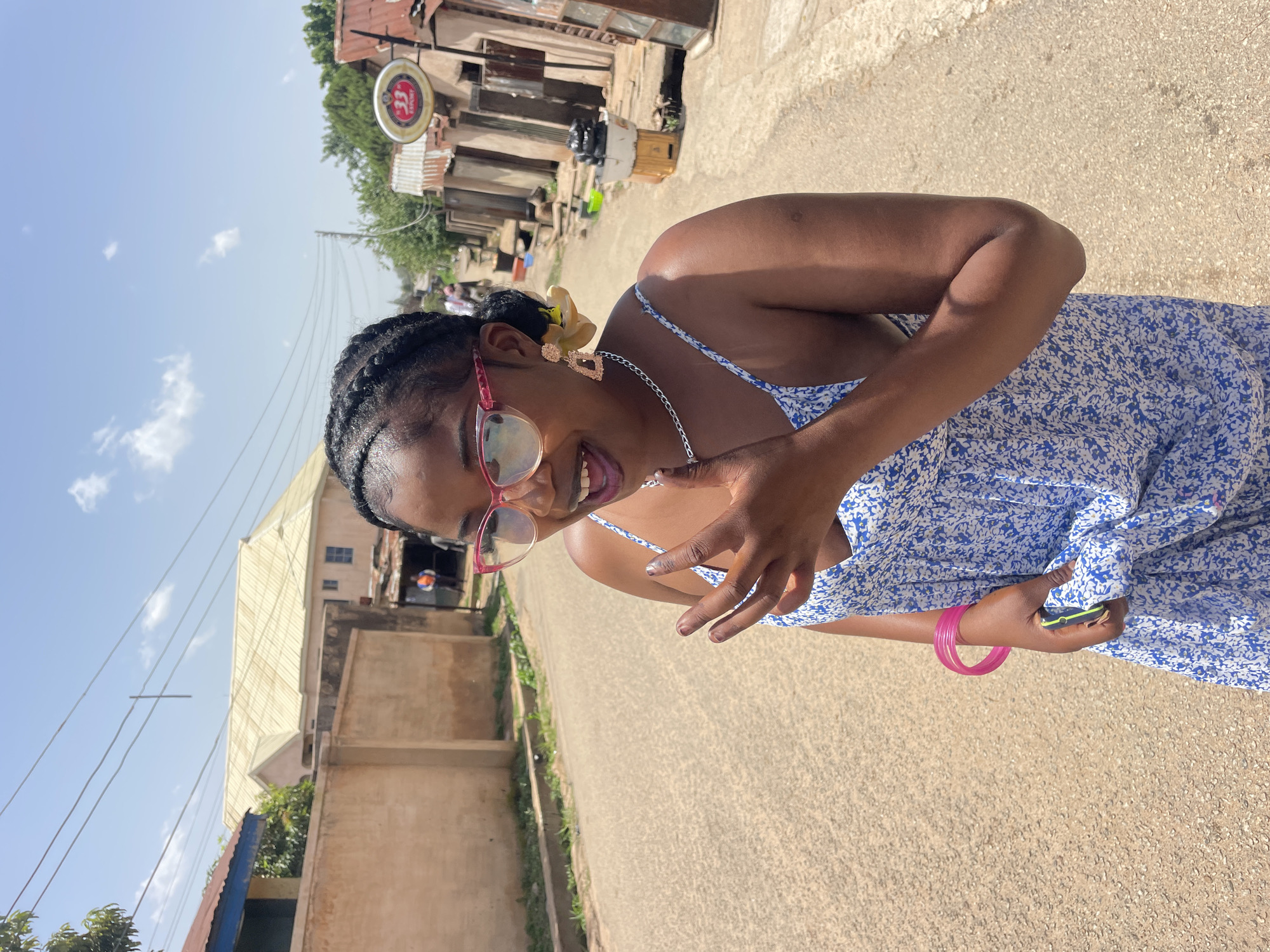 A young woman is standing on a dirt road in what appears to be a residential area in Jos, Nigeria. She is wearing a blue and white floral print sundress, pink-rimmed glasses, a silver necklace, and large, dangling earrings. Her hair is styled in braids. She is smiling with her mouth open and her right hand is raised near her face, with her fingers extended. She is holding a neon yellow cell phone in her left hand, and she has a pink bracelet on her left wrist. The road is paved with dirt and gravel, and there is a concrete wall on the left side of the image. In the background, there are several buildings with corrugated metal roofs, some of which appear to be shops or homes. There are also trees and power lines visible. The sky is blue with some scattered clouds.