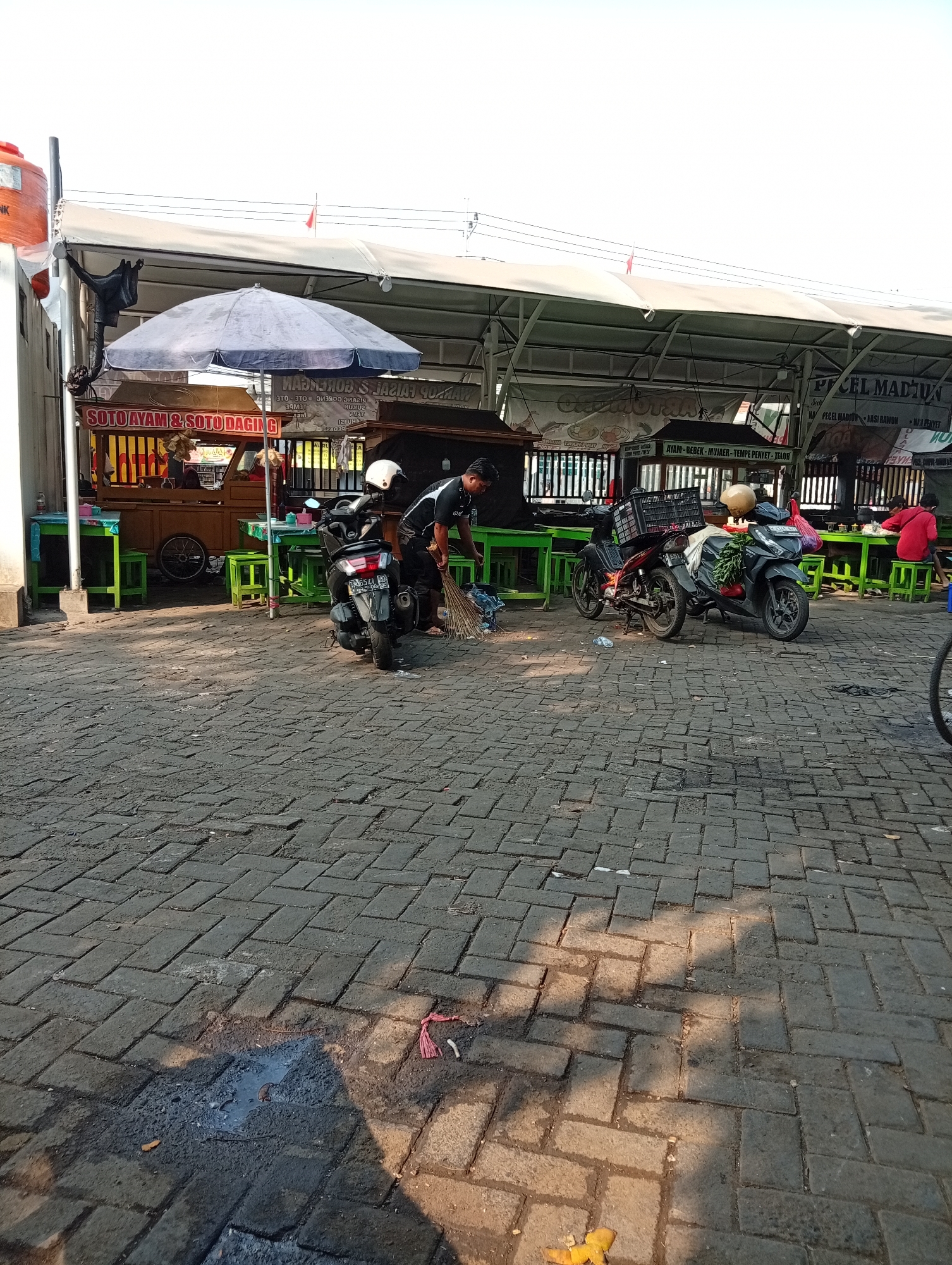 The image captures an outdoor scene in the city of Larangan, Indonesia, likely a marketplace or a food stall area. A man is seen sweeping the cobblestone ground with a broom. Several motorcycles are parked on the pavement, some with baskets or bags attached. In the background, there are food stalls with colorful awnings, displaying menus with Indonesian text like "SOTO AYAM & SOTO DAGING" and "PECEL MADIUN." Green tables and stools are set up for seating. The overall environment appears to be busy, with several people present, some seated and others standing. Sunlight casts shadows on the ground, indicating it is daytime. The scene is set against a backdrop of simple structures and overhead coverings typical of an open-air commercial setting.