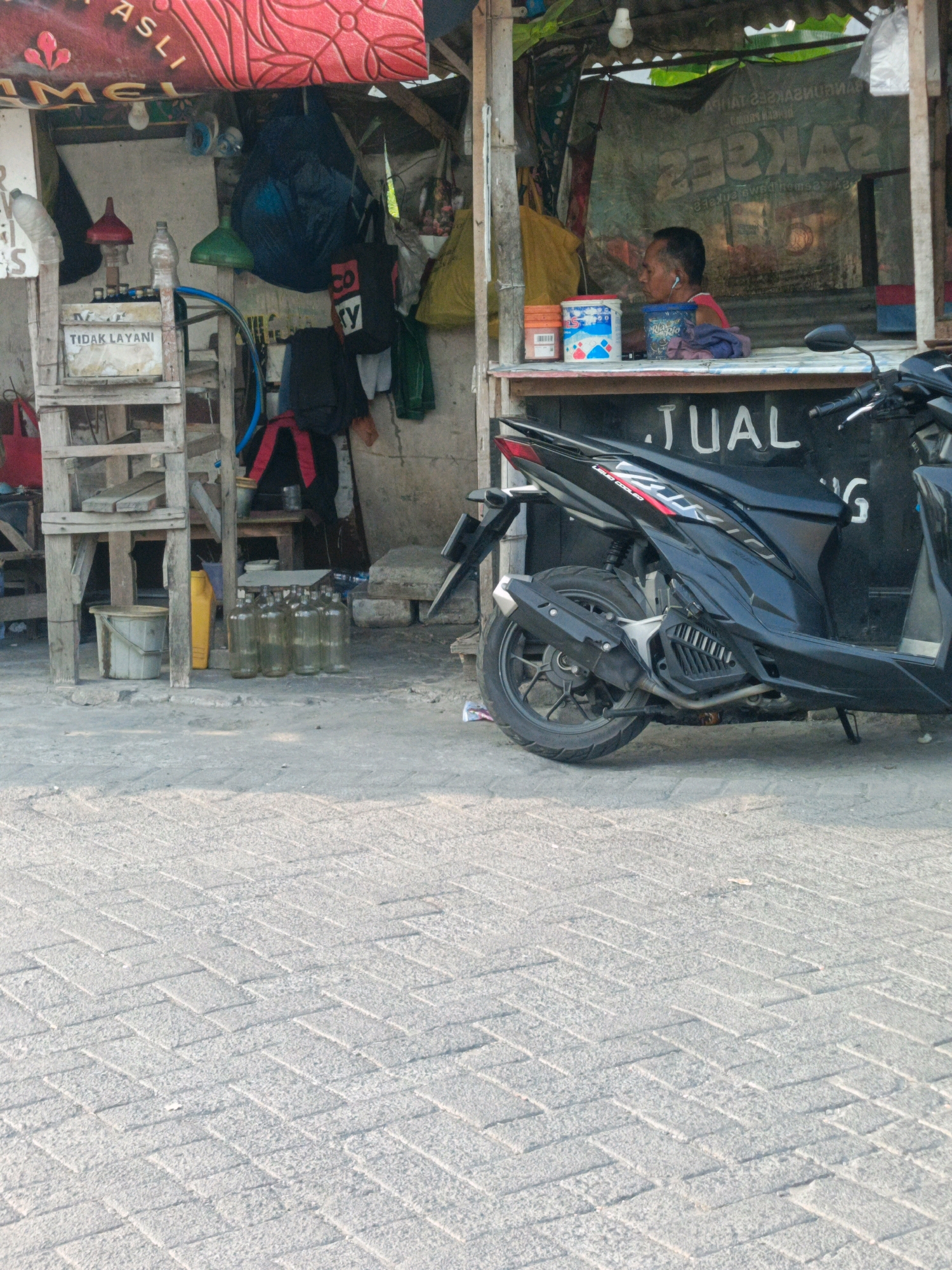 The image depicts a street scene in Pepe, Indonesia, likely a small business or roadside stall. In the foreground, a black motorcycle is parked. Behind the motorcycle, a stall is visible with a sign that reads "JUAL" (meaning "FOR SALE" in Indonesian). A man is seated inside the stall, wearing earphones. The stall appears to be selling various items, including bottles, buckets, and possibly fuel. To the left, a wooden structure holds more bottles and a yellow container. The ground is paved with interlocking bricks. The overall impression is of a busy, utilitarian street environment.