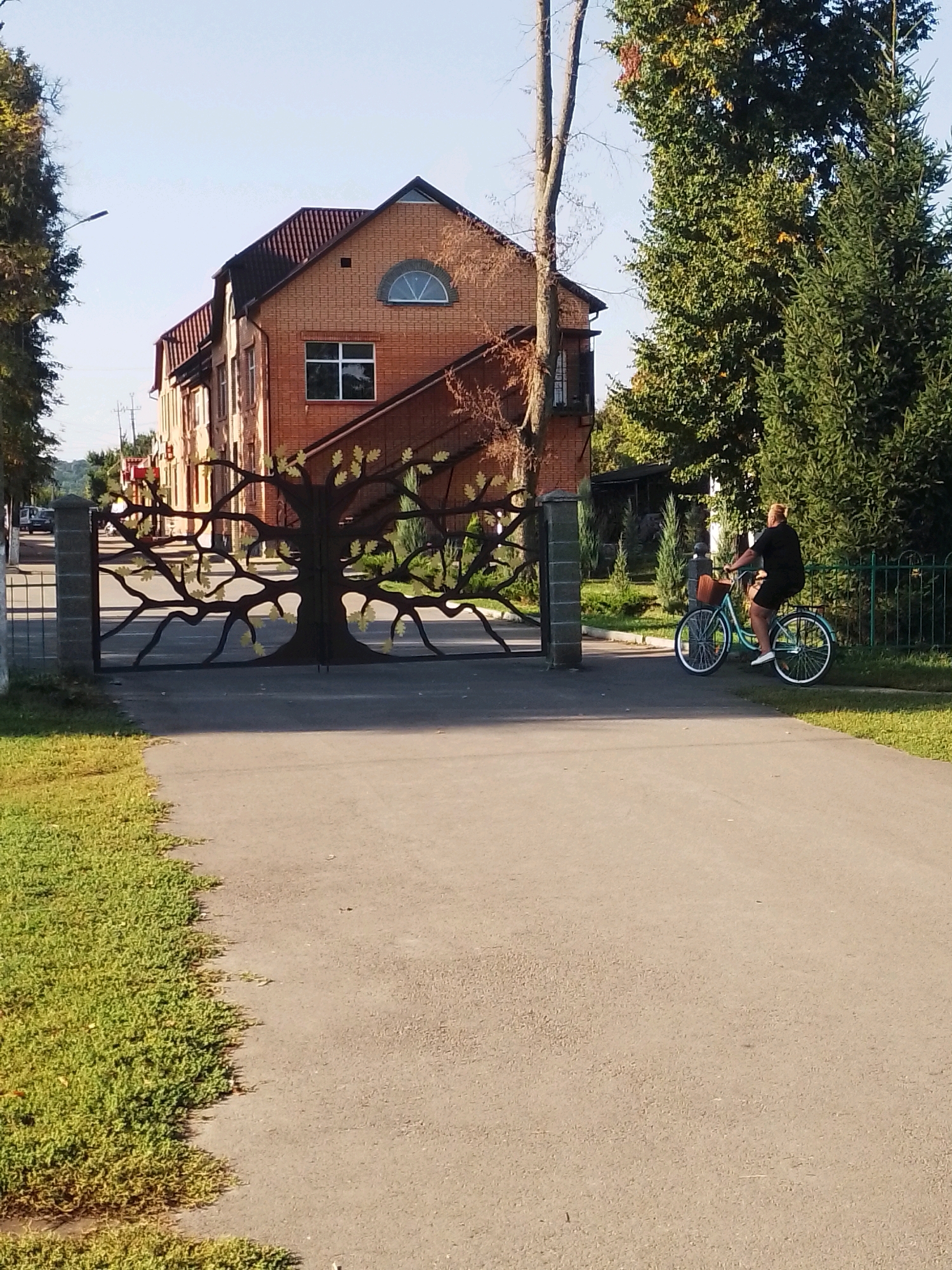 A person is riding a bicycle on a paved road in front of a brick building with a decorative metal gate featuring a tree motif. The bicycle, light blue in color, has a woven basket on the front. The person is wearing a black outfit and white shoes. The building, likely in Velyka Bahachka, Ukraine, has a red-tiled roof and several windows, one of which is arched. Adjacent to the road is a grassy area, and further back, there are trees and a green metal fence. The sky is clear and blue, indicating a daytime scene.
