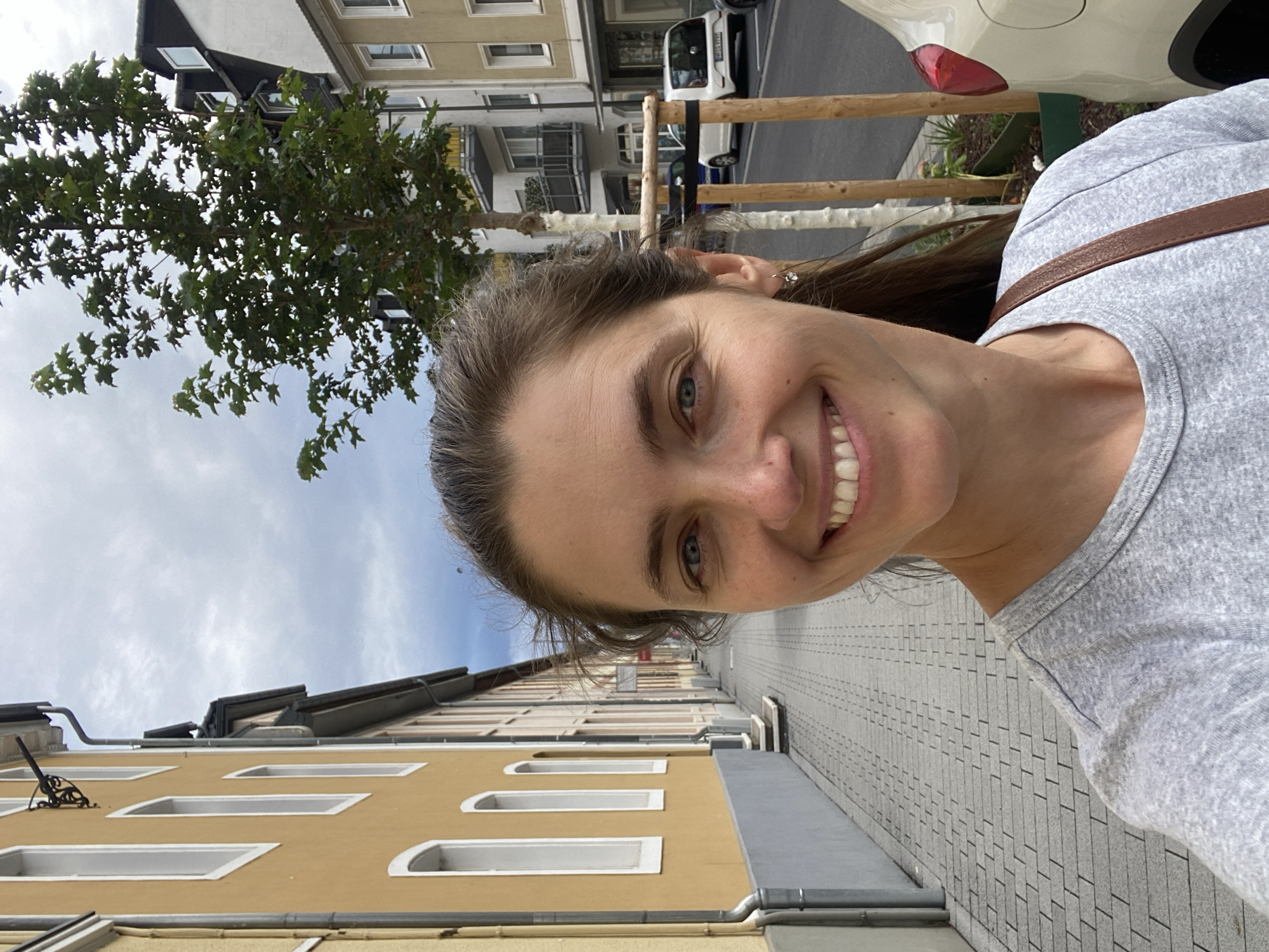 A smiling woman is taking a selfie on a sidewalk in front of buildings and a street in Neuwied, Germany. The woman has brown hair pulled back, blue eyes, and is wearing a grey t-shirt and a brown strap across her shoulder. In the background, there are yellow and white buildings, a paved sidewalk, a street with parked cars, and a tree. The sky is overcast.