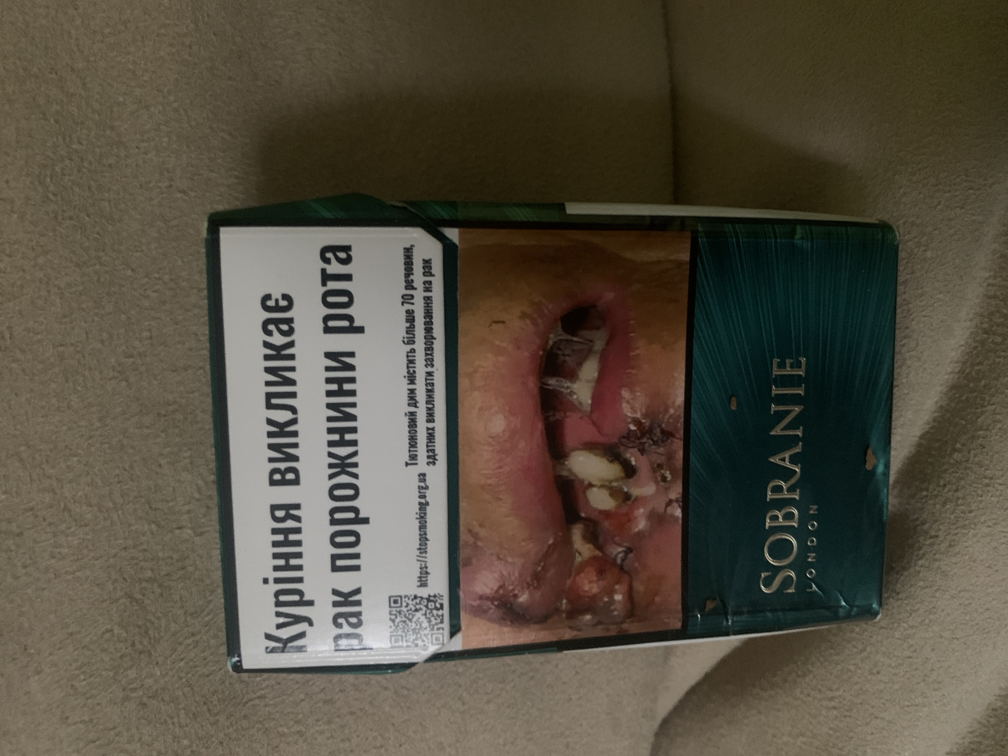 This image shows a close-up of a pack of Sobranie cigarettes. The pack features a graphic warning image of diseased oral tissue and text in Ukrainian that translates to "Smoking causes mouth cancer." Below the warning, the brand name "SOBRANIE" and "LONDON" are visible in gold lettering on a dark teal background. The pack appears to be placed on a textured surface, possibly a couch or carpet, in an indoor setting. There is no visible indication of the location being Skarydzew, Poland.