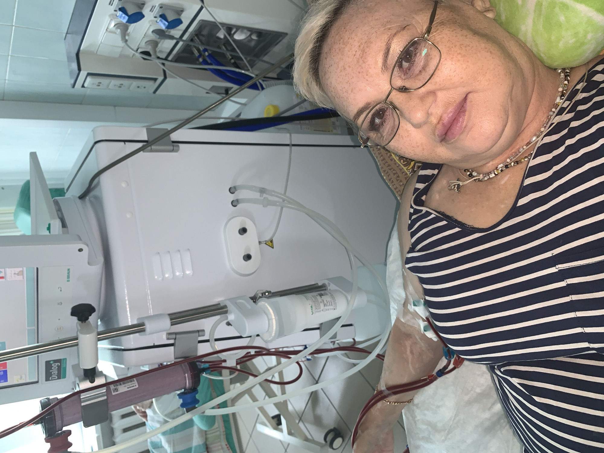 A woman is lying in a hospital bed, undergoing dialysis. She is wearing a striped shirt and glasses. A dialysis machine, with tubes and filters, is visible next to the bed. The room appears to be a medical facility, with tiled walls and medical equipment. The location is identified as Chaikovsky, Russia, although no specific landmarks confirm this.