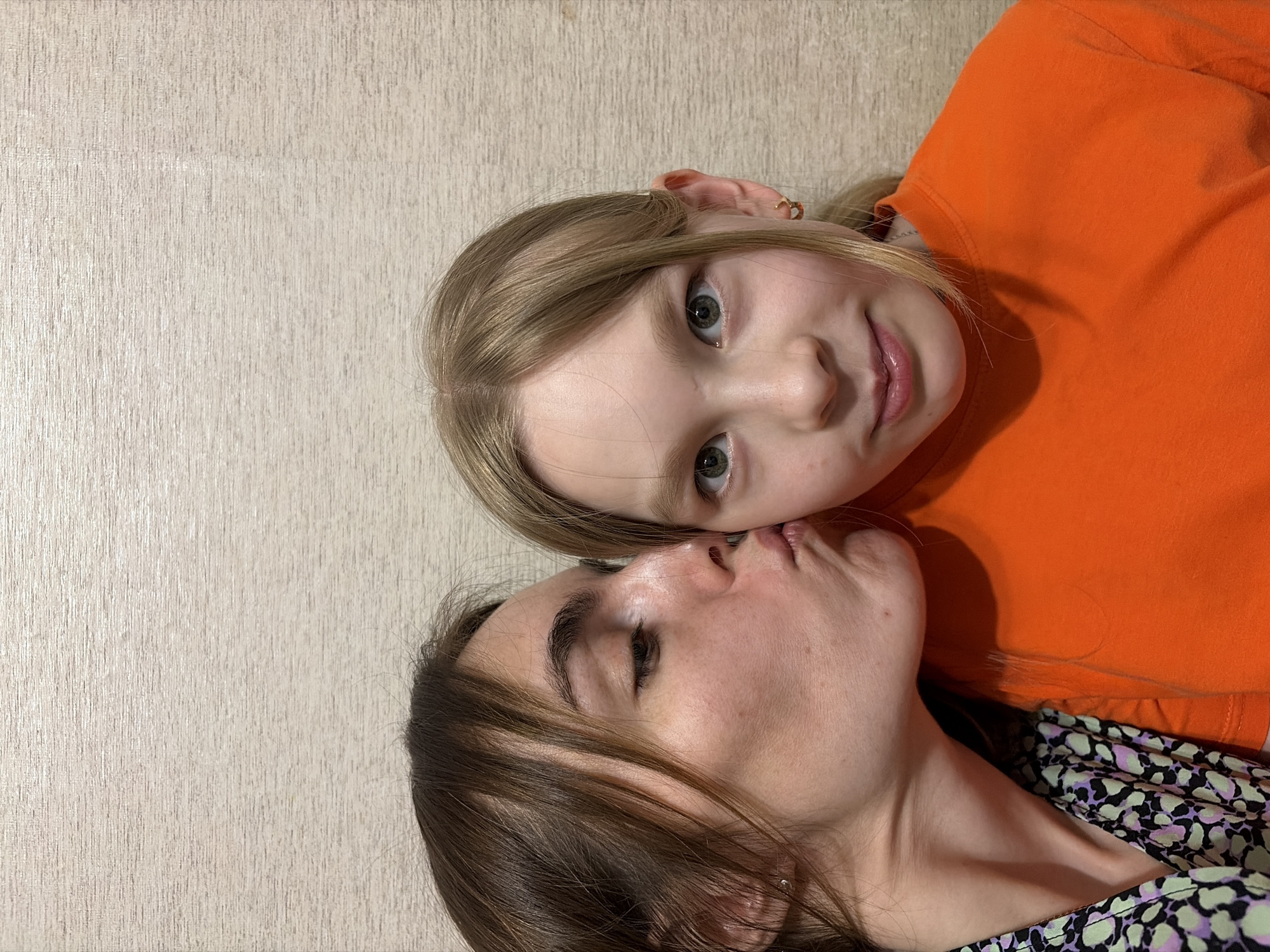 The image features a fair-skinned woman and a young girl, presumed to be mother and daughter, lying close together. The girl, dressed in an orange shirt, has blonde hair and light-colored eyes. The woman, wearing a patterned shirt, has brown hair and her eyes are closed. The background appears to be a textured wall, possibly wallpaper. There is no indication in the image to suggest a location in Izhevsk, Russia. The interaction suggests affection or intimacy.