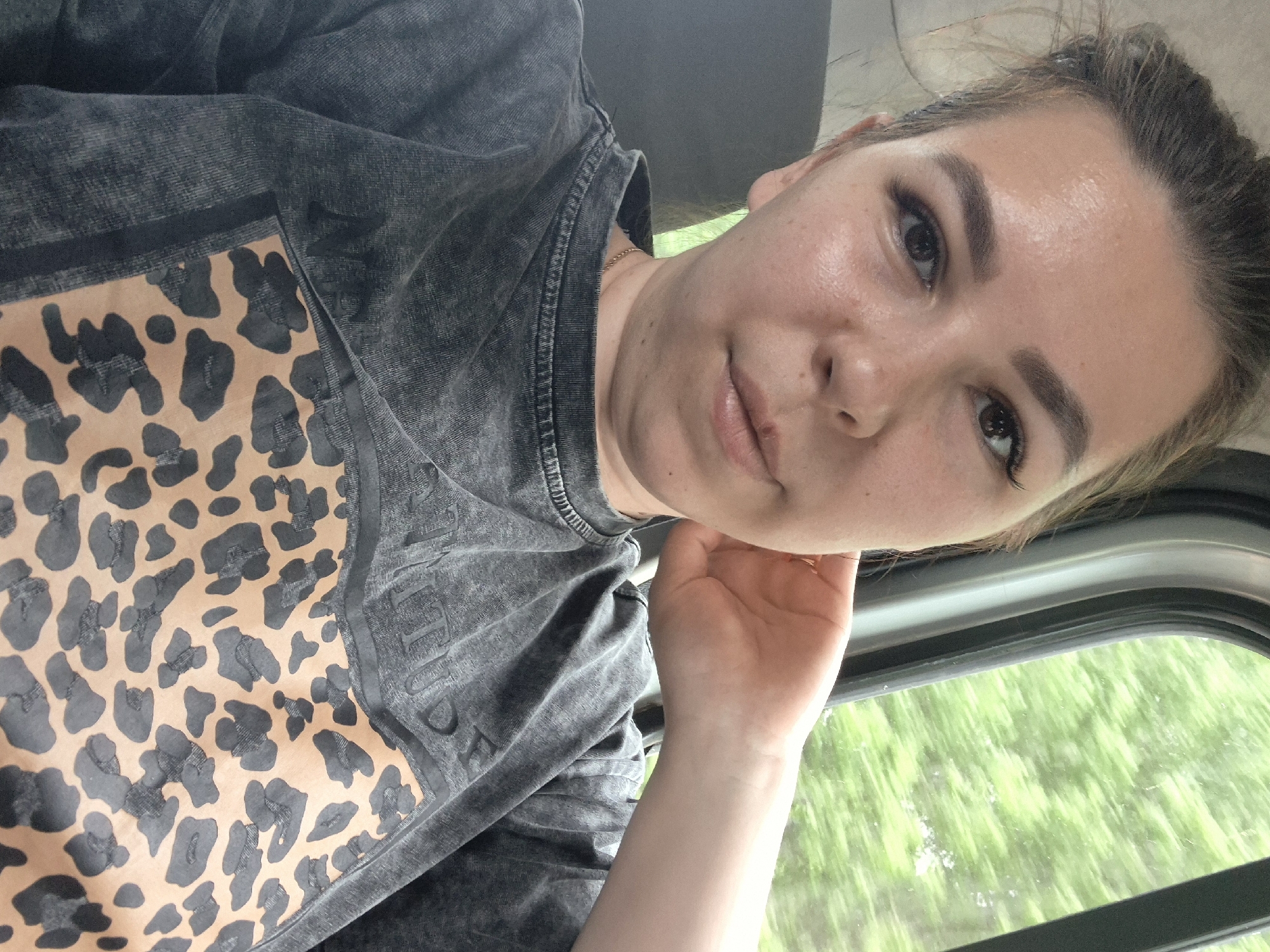 A young woman is seated inside a vehicle. She is wearing a grey t-shirt with a leopard print design on the left side. She is holding her face with her right hand, resting her elbow on the windowsill. Through the window, blurred green trees are visible.