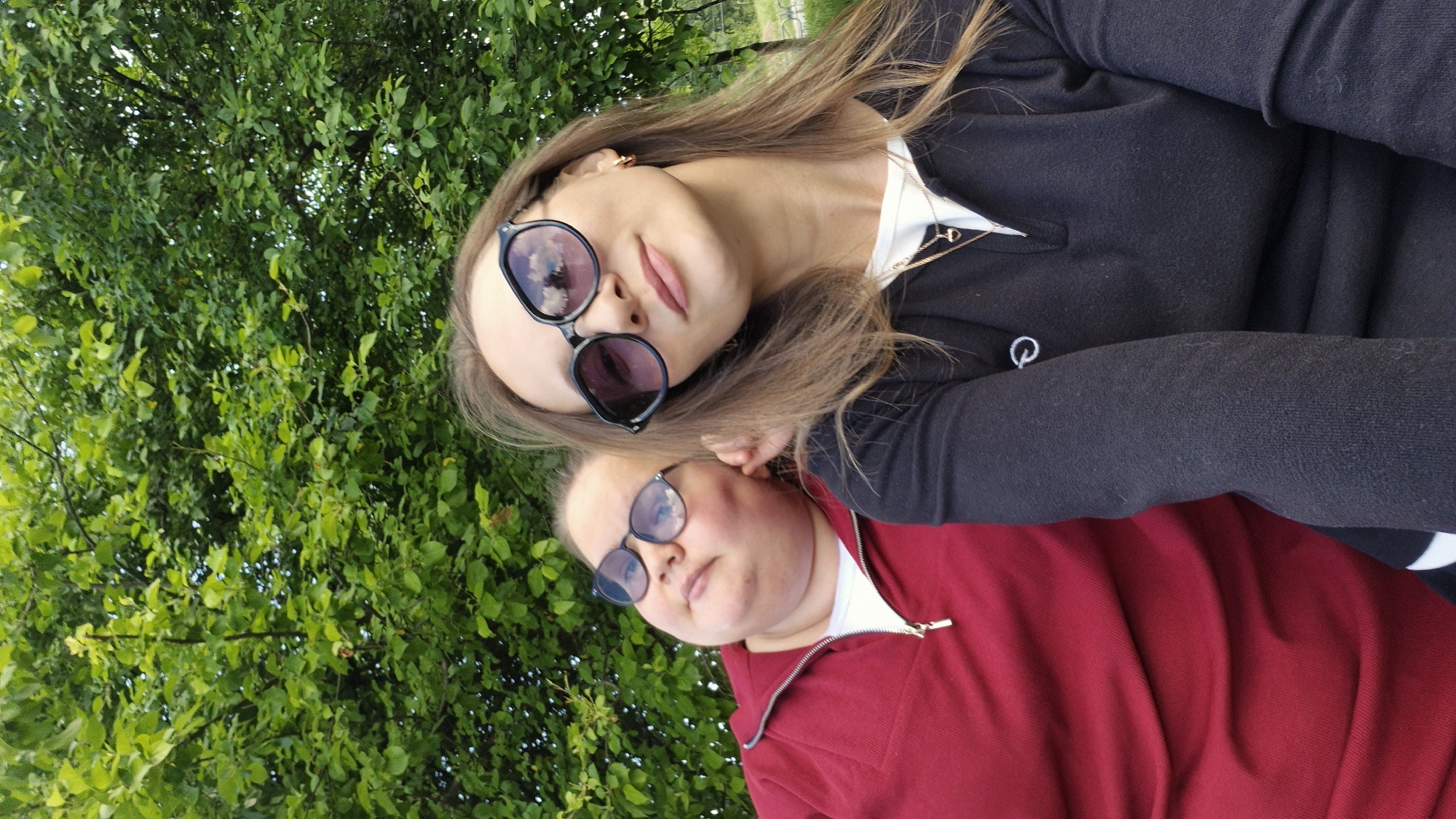 Two women pose for a selfie against a backdrop of lush green foliage. The woman in the foreground wears a red zip-up sweatshirt and black-framed glasses. The woman behind her has long brown hair and is wearing a dark-colored sweater, a white shirt, and sunglasses. The selfie was taken in Chaikovsky, Russia, as reflected in the sunglasses.