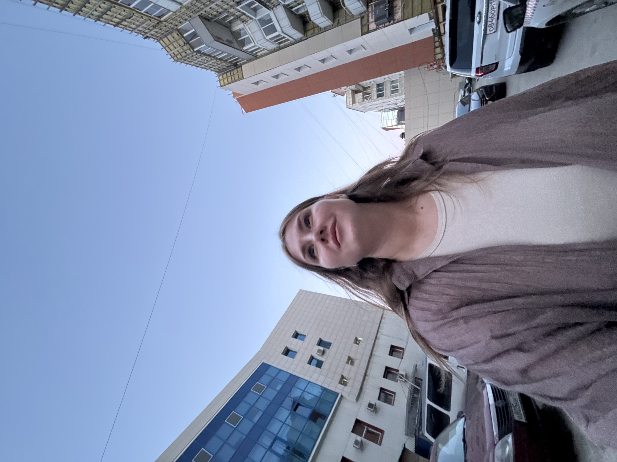 A low-angle outdoor shot captures a woman from a close distance. She is wearing a light-colored top and a brown cardigan. Buildings rise in the background, including one with a modern blue glass facade. A white vehicle is visible, bearing the license plate "0846OP159." The location is Perm, Russia.