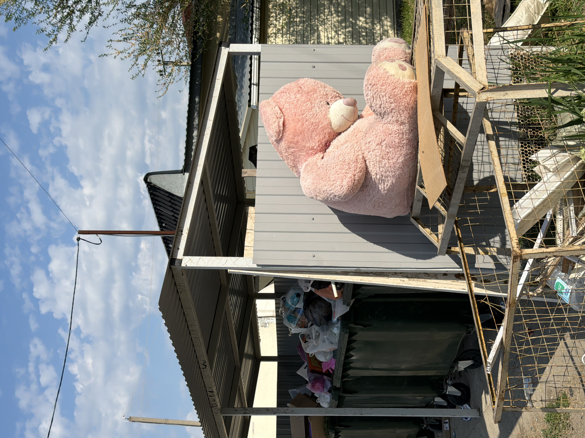 A large, pink teddy bear is leaning against the grey wall of a small, dilapidated building. The building appears to be an outbuilding, possibly a shed or storage area, judging by the cluttered interior visible beneath the overhang. The area around the building shows signs of disrepair and neglect, with an old, rusty metal structure nearby. It is filled with refuse. The location is Leningradskaya, Russia. The sky is visible, featuring white clouds and blue sky.