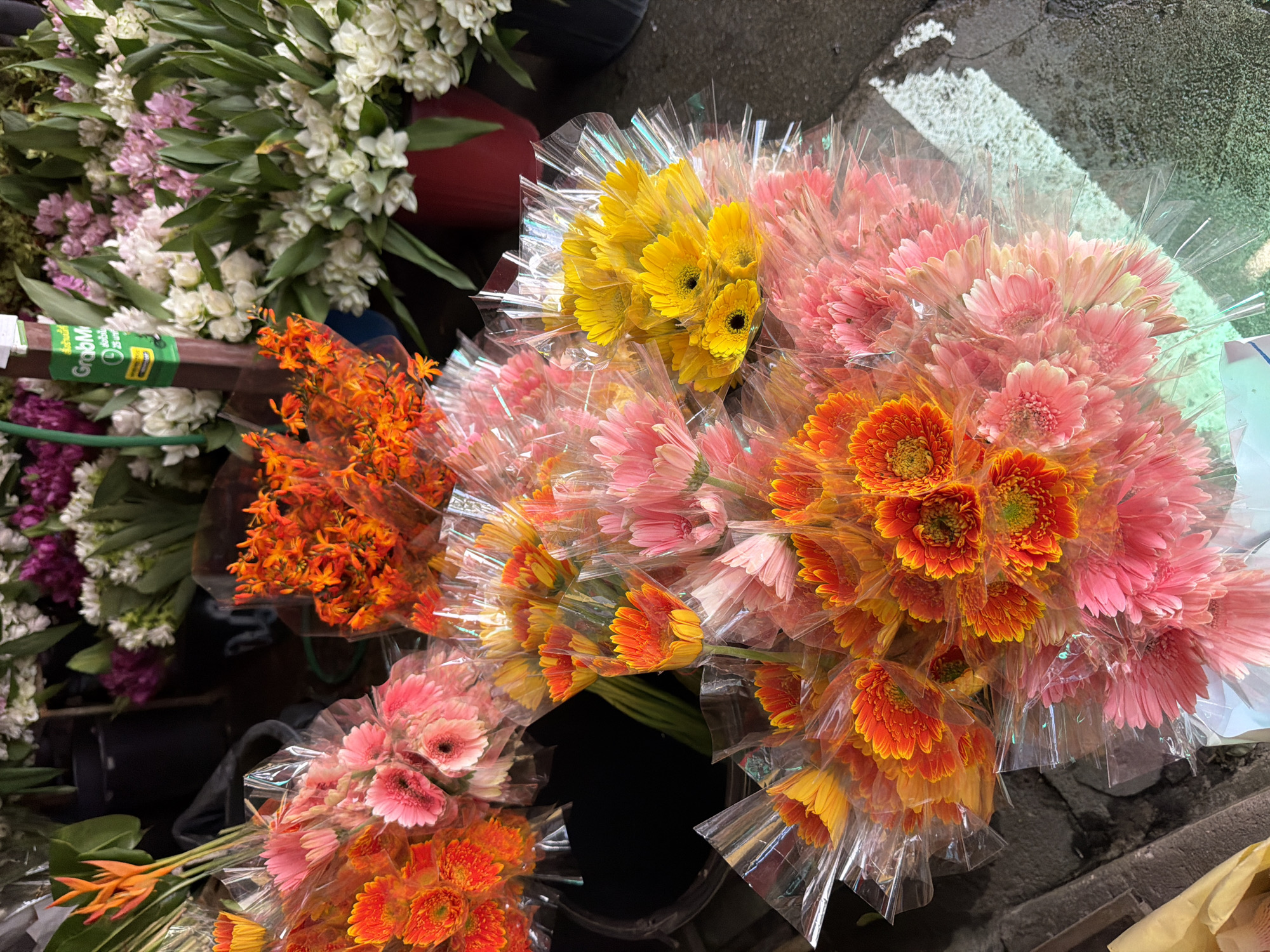 The image depicts a vibrant flower stall, likely located in the city of Chiang Mai, Thailand. The foreground is dominated by several bouquets of gerbera daisies, neatly wrapped in clear plastic. These flowers are a riot of color, featuring shades of bright orange, yellow, and soft pink. Some of the orange gerberas are a deeper, fiery hue, with yellow centers. The pink gerberas are a softer, more pastel shade. Behind these primary bouquets, more flowers can be seen, including a cluster of smaller, bright orange flowers, possibly orchids or a similar variety. In the background, larger arrangements of white and pink flowers, possibly a type of ginger or other tropical bloom, are visible, some in a dark red pot. The stall appears to be outdoors, with a rough, textured surface that could be a sidewalk or pavement. A green sign with Thai script and the Grab logo is partially visible, indicating the presence of a delivery service or perhaps a vendor accepting online orders. The overall impression is one of abundance and a lively marketplace setting.