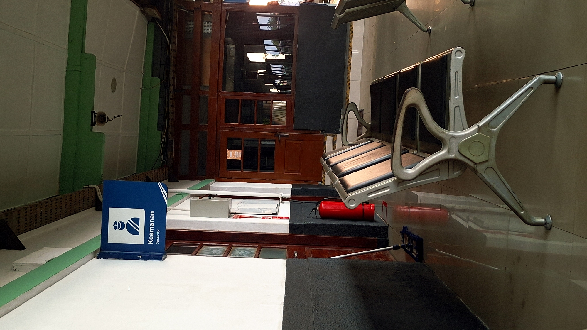 The image depicts an interior space, likely a train station or similar transit hub, in Karawang, Indonesia. The foreground features rows of metal and wood waiting benches affixed to a tiled wall. In the midground, a blue sign with an icon and the words "Keamanan Security" (security in Indonesian) sits on a white surface. Behind this is a brown wooden structure resembling a partitioned door or window frame. A red fire extinguisher is positioned near the benches. In the background, there is a glimpse of an outdoor area through the wooden structure, indicating a potential entrance or exit. A mop is visible, suggesting recent or ongoing cleaning activity. The overall scene is one of a functional public space.