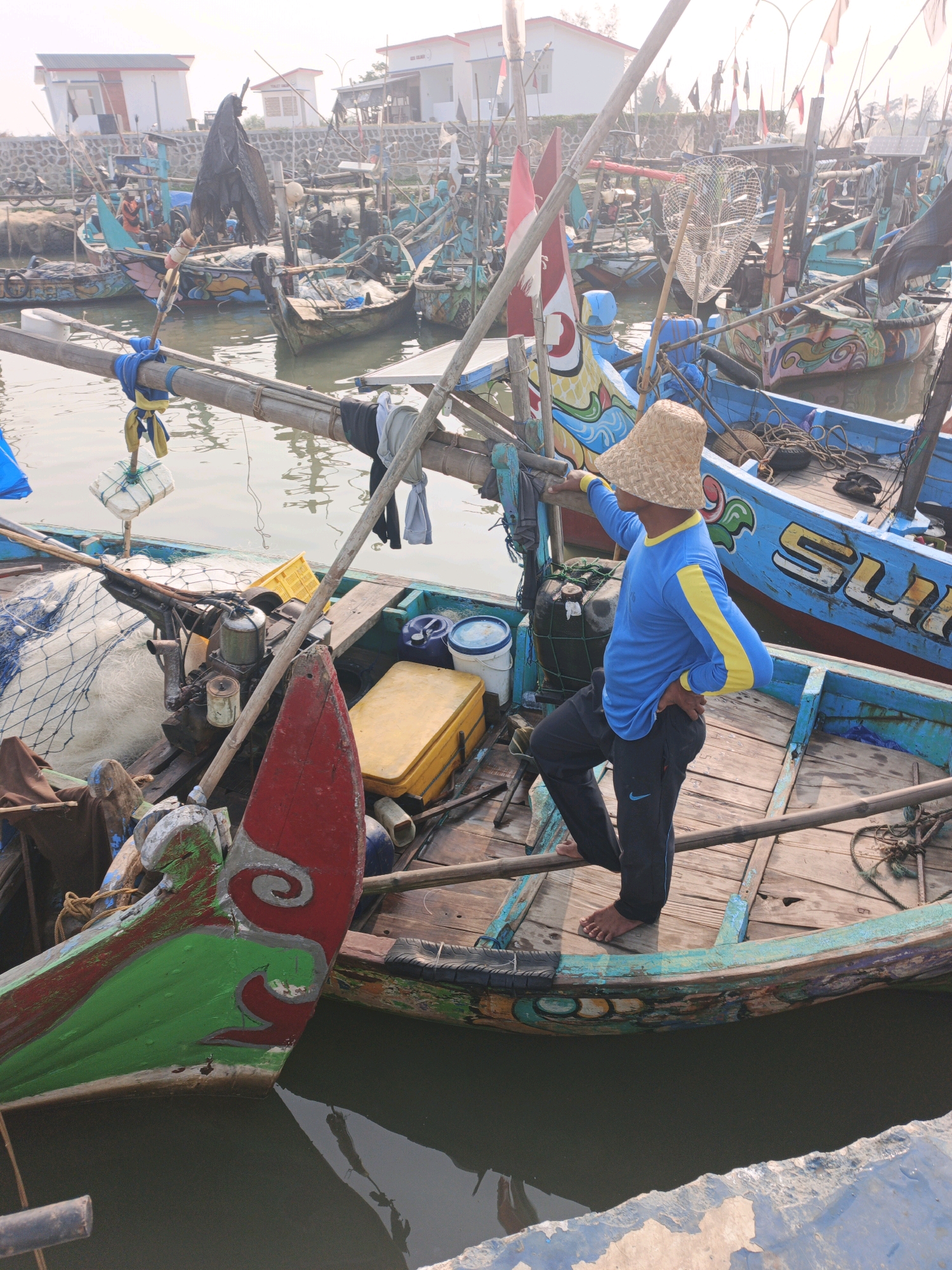 A man wearing a straw hat, a blue long-sleeved shirt with yellow accents, and black pants stands on a wooden boat. His left hand is on his hip, and his right arm is extended, resting on the boat's railing. The boat is docked among many other similar boats in a harbor. The boats are painted in various bright colors, with intricate designs. Fishing nets, engines, and storage containers are visible on the boats. In the background, there are buildings and flags. The scene appears to be a fishing village in Indonesia.