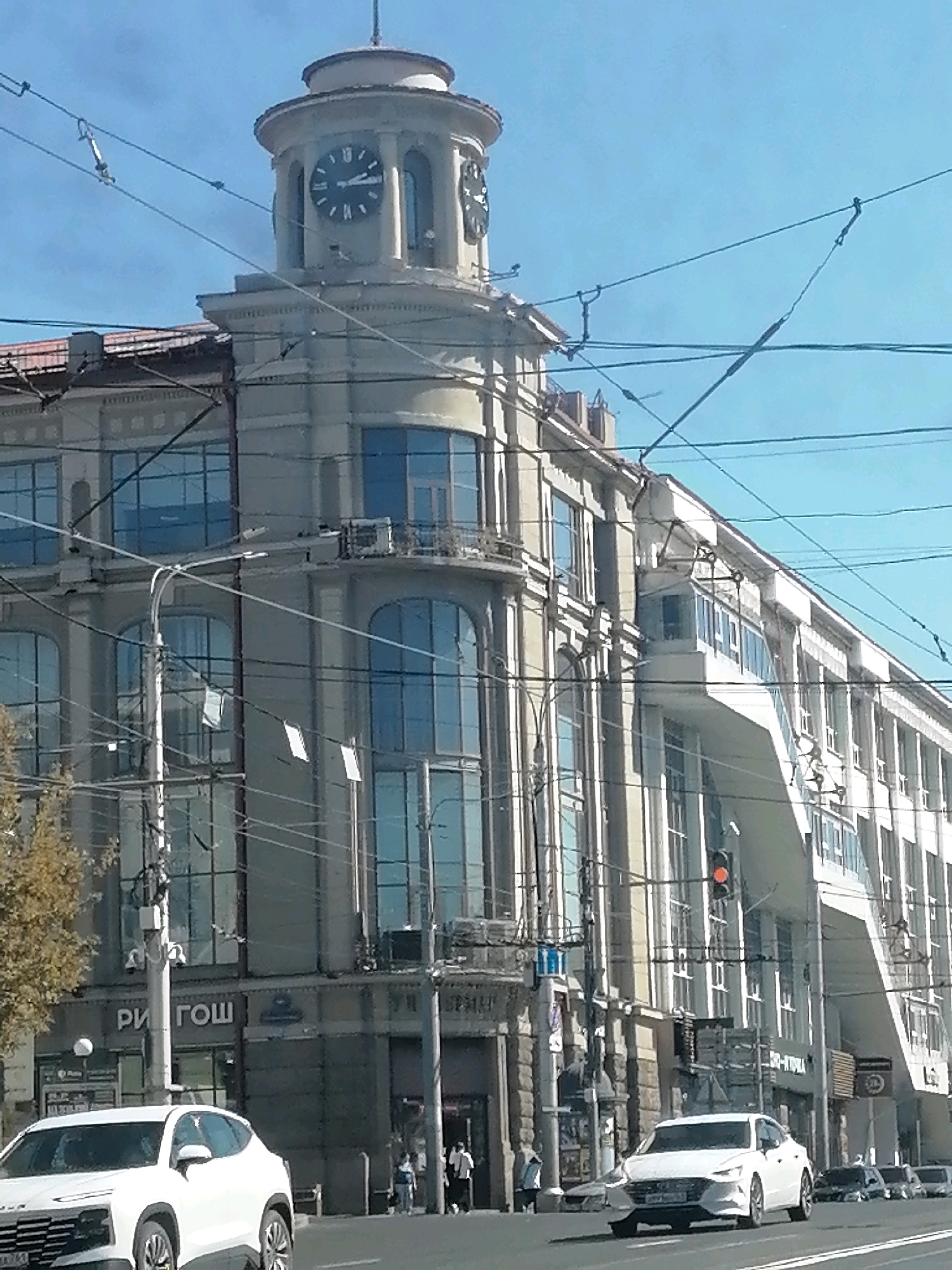 This is a daytime, eye-level shot of a large, multi-story building in Rostov-on-Don, Russia, featuring a prominent clock tower. The building has a light-colored facade with a classical architectural style, characterized by large, arched windows and decorative cornices. The clock tower has a circular base and a domed top, with Roman numerals marking the time on its face. Overhead wires, likely for public transportation, crisscross the scene.
In the foreground, a white car is driving on a street. Further down the street, other vehicles are visible, as well as pedestrians walking on the sidewalk. A sign with Cyrillic lettering, reading "РИГОШ", is visible on the lower part of the building. The sky is clear and blue. The overall impression is of a busy urban environment with a mix of old and new architecture.