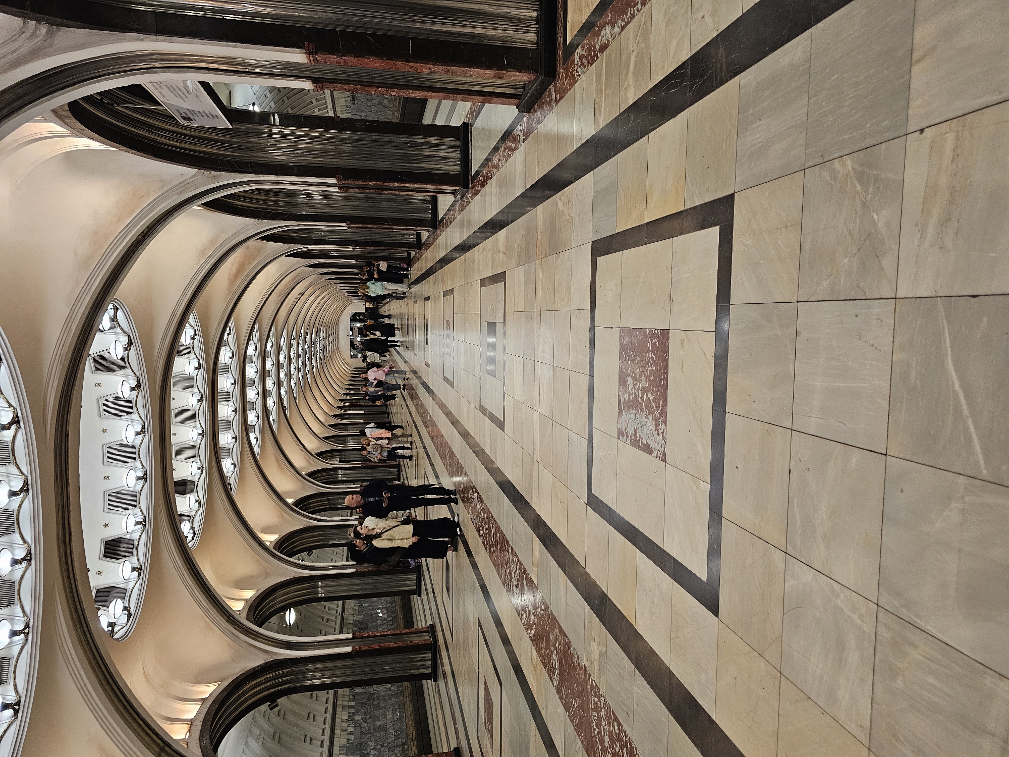 This image depicts the interior of a Moscow Metro station, likely a grand, ornate one, possibly in the style of Stalinist architecture, characterized by its high, vaulted ceilings with intricate lighting fixtures and extensive use of marble and polished stone. The station features a long, wide concourse lined with archways supported by decorative columns. The floor is paved with large rectangular tiles in a pattern of light beige, cream, and darker grey or black strips, with some square inlays featuring reddish-brown veined marble.
Several individuals are visible walking along the concourse. In the foreground, on the left side, a group of four people are gathered, appearing to be conversing. Three of them are women, one wearing a light-colored jacket and dark trousers, another in a dark outfit, and a third in a light-colored blouse and dark trousers. A man in a dark suit and tie stands with them. Further down the concourse, more people are seen walking in both directions, some appearing to be tourists due to their attire and the presence of cameras. The lighting is bright and even, emanating from the decorative fixtures along the ceiling. The overall impression is one of grandeur, spaciousness, and public transit functionality.