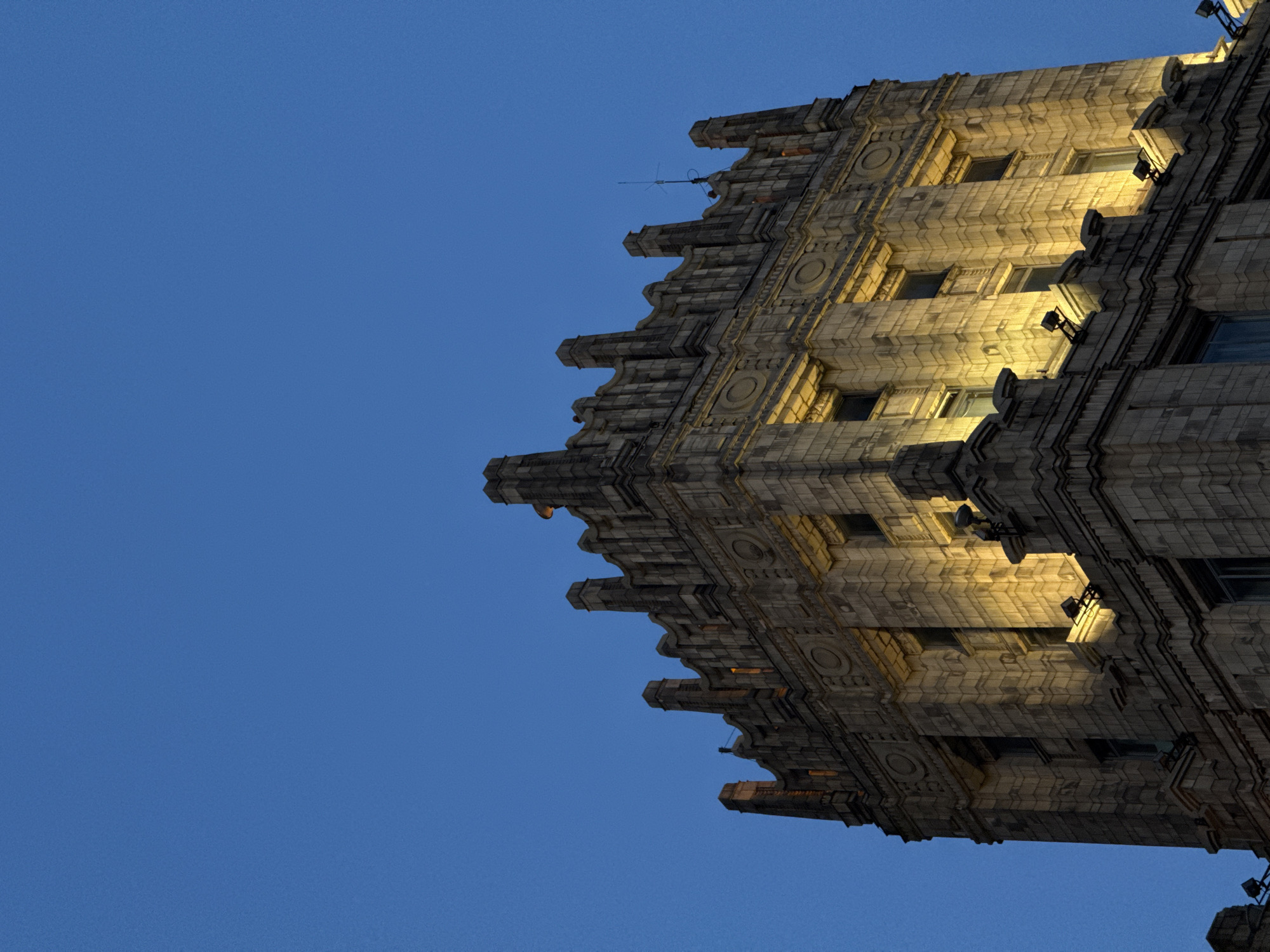 The image shows a building's corner facade, likely located in Moscow, Russia, given its architectural style. The building is constructed with light-colored stone blocks featuring decorative elements like circular details and ribbed patterns. The facade is illuminated by warm, artificial light, creating contrast and highlighting the architectural details against the dark blue evening sky. The building appears to be an example of the Stalinist architecture era, common in Moscow, Russia. An antenna is present on the roof.