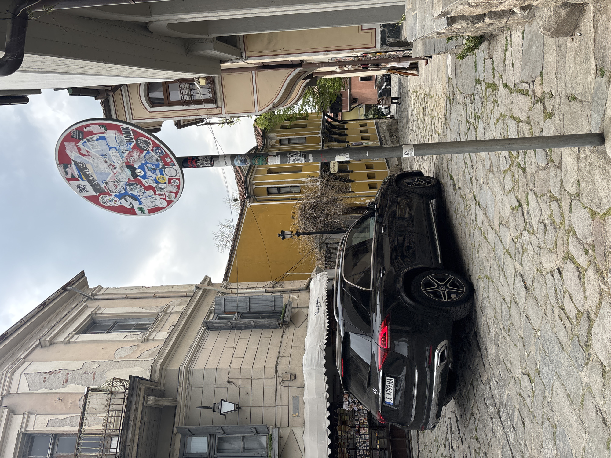 The image depicts a narrow street in Plovdiv, Bulgaria, paved with cobblestones. A black car is parked on the right side of the street. A metal pole is situated near the car, supporting a circular sign covered with stickers. The buildings on either side of the street are old, with visible signs of wear and tear. One building has a partially open awning. In the distance, two figures stand near another building. The sky is overcast.