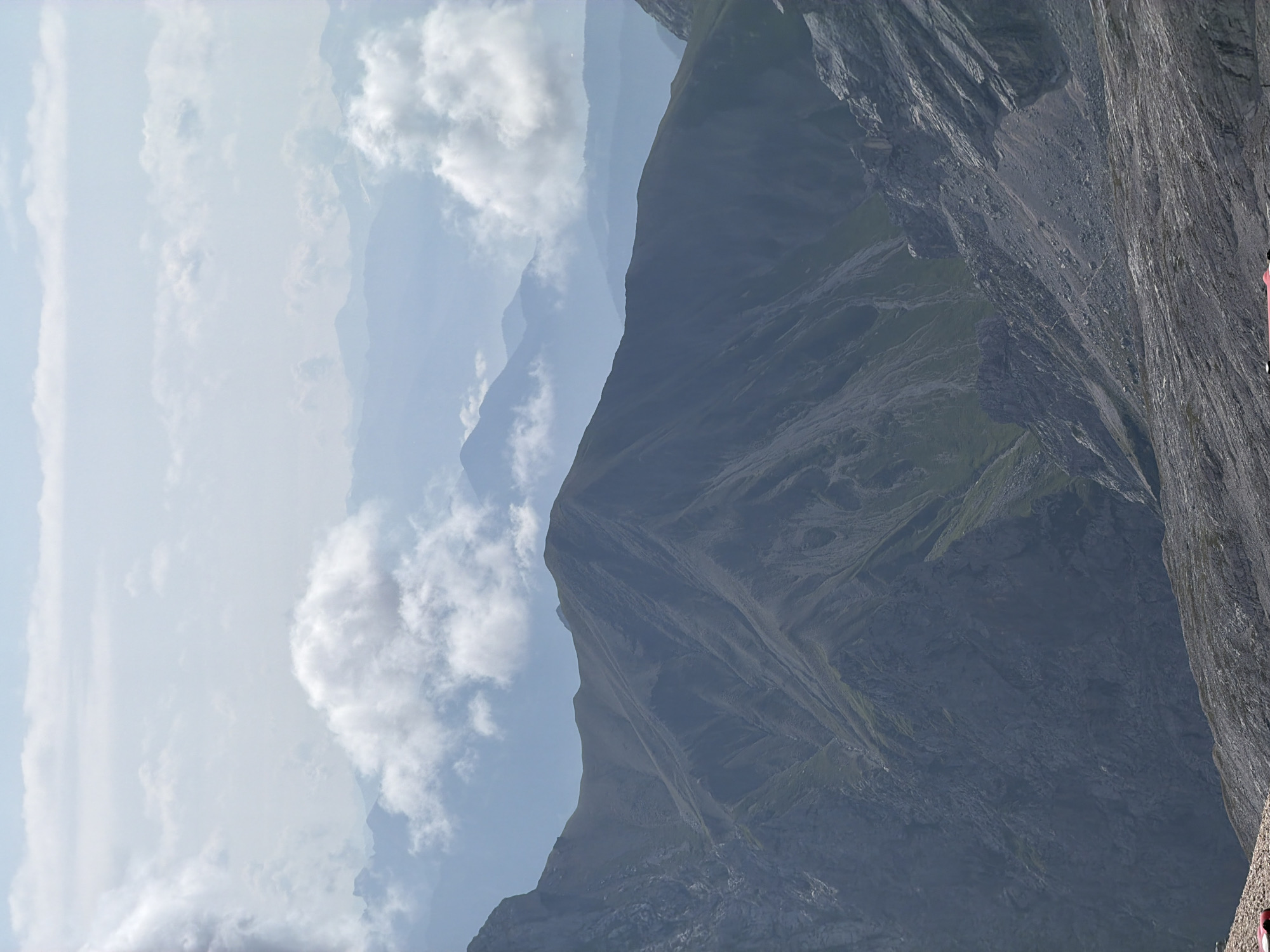 The image depicts a mountainous landscape, likely in the Alps, with layers of peaks fading into the distance under a hazy sky. Clouds are present in the sky and partially obscure some of the further mountain ranges. The foreground shows rocky terrain and steep slopes with some sparse green vegetation. There are no visible people or specific city landmarks that would definitively place it in Garmisch-Partenkirchen, Germany, although the scenery is consistent with the region's topography. A small section of a red object, possibly a building or vehicle, is visible in the lower left corner of the image.