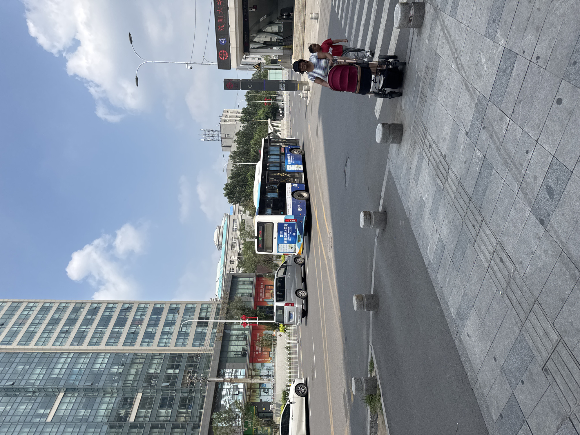 The image captures a street scene in Dadong District, China, on a clear day with scattered clouds. In the foreground, a grey paved sidewalk with tactile paving leads to a crosswalk. Several cylindrical concrete bollards are placed along the edge of the road.
On the road, a silver minivan is parked in the middle lane, and a white car is to its left, turning. A blue and white city bus is approaching the crosswalk from the right. A woman wearing a striped shirt and a hat is pushing a red stroller across the crosswalk, accompanied by a young boy in a red shirt.
In the background, a tall modern building with a glass facade stands on the left, reflecting the sky and clouds. Across the street, there are other buildings, trees, and streetlights. A subway station entrance with signage indicating "沈阳大学" (Shenyang University) is visible on the right side of the image. The overall impression is that of a busy urban environment during daylight hours.