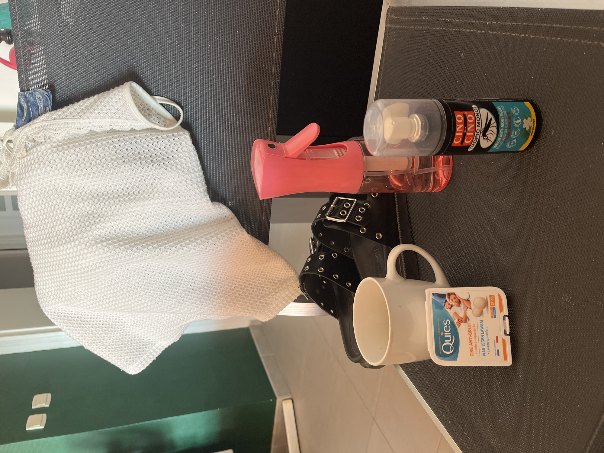The image depicts a still life composition on a dark, textured surface, possibly a table or chair, indoors. In the foreground, a white mug is positioned to the left, with a small white package labeled "Quies" resting in front of it. The Quies package features an illustration of a couple sleeping and text in French and Dutch, indicating earplugs. To the right of the mug, a black pair of sandals with multiple silver buckles and eyelets are partially visible. Behind the mug and sandals, a pink spray bottle with a clear liquid is present. To the right of the pink bottle, a black aerosol can with a white cap is standing upright. The can has a label with prominent red and white text "CINO CINO", and a black mosquito illustration, suggesting it is insect repellent. In the background, a white waffle-knit fabric, possibly a towel or clothing item, is draped over something dark, likely the back of a chair. Two light switches are visible on a dark green wall to the far left. The overall scene is well-lit, with natural light appearing to come from the left. No people are present in the image. The location is identified as Palavas-les-Flots, France.