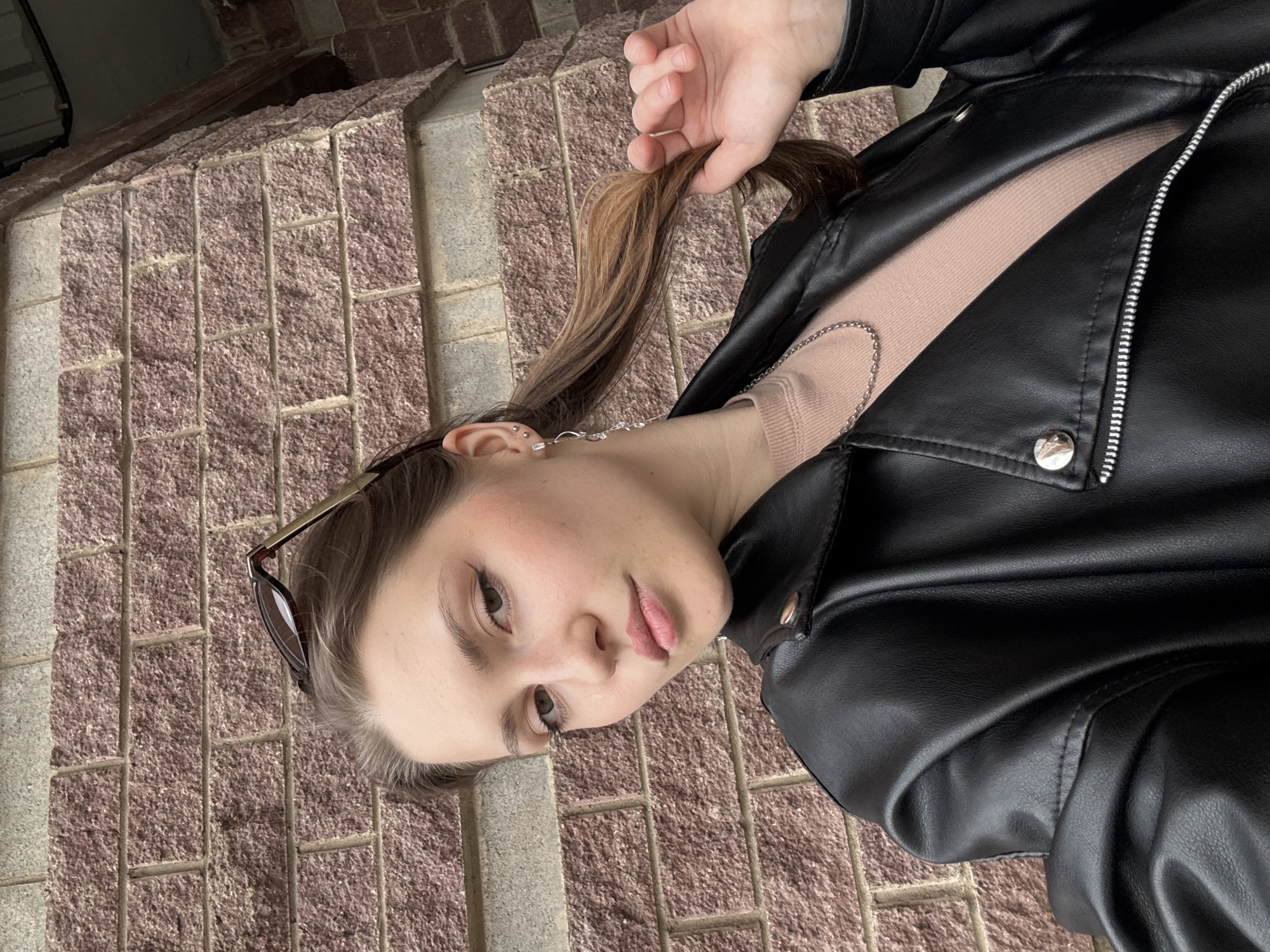 This image is a horizontally oriented portrait of a young person, likely female, captured from the chest up. The subject has fair skin, medium brown hair, and dark eyebrows. She is looking directly at the camera with a neutral to slightly contemplative expression, her lips softly parted. She wears a black faux leather jacket with a prominent silver zipper and snap buttons, over a light pink or beige ribbed top. A delicate silver chain necklace is visible around her neck, and she has multiple silver earrings on her left ear, including a longer dangling one. Sunglasses with dark lenses are perched on her head. Her right hand is gently holding a section of her hair near her head.
The background is a reddish-brown textured brick or stone wall with lighter mortar lines, suggesting an outdoor or urban setting. The lighting appears even, indicating daytime. The overall impression is a casual portrait. The image is located in Saint Petersburg, Russia. There is no visible text.