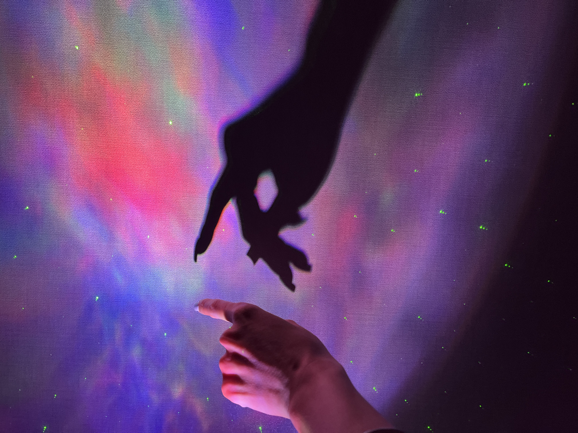 The image depicts a person's hand, illuminated by pink light and casting a shadow, pointing upwards against a backdrop of a nebula-like projection in vibrant purple, pink, and blue hues, interspersed with small green dots resembling stars. The projection creates a textured, canvas-like surface. The shadow of another hand, with fingers splayed, is visible to the right of the projected nebula. There is no indication of the city of Nizhny Novgorod, Russia, or any specific urban location. The scene appears to be staged or part of an art installation, focusing on light and shadow play.