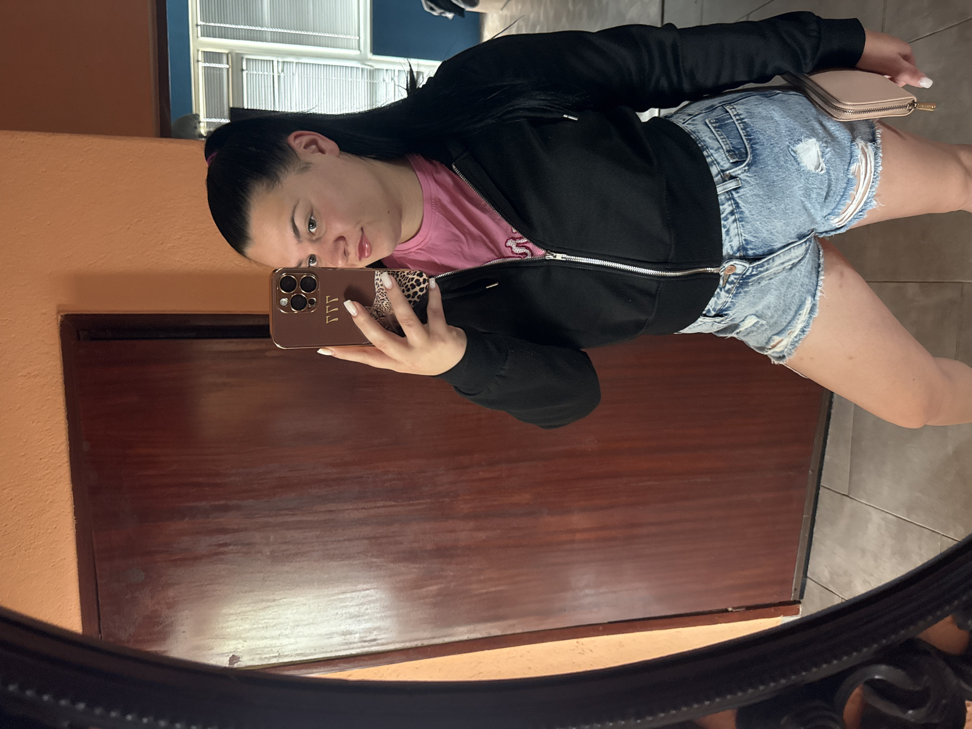 A young woman with black hair pulled into a ponytail is taking a mirror selfie in what appears to be a home interior. She is wearing a pink t-shirt, a black zip-up hoodie, and distressed denim shorts. She is holding a smartphone with a leopard print phone case. A small pink wallet is visible in her right hand. The background shows a wooden door, an orange wall, and tiled flooring. The scene does not provide any specific indicators of the city of Rogosh, Bulgaria.