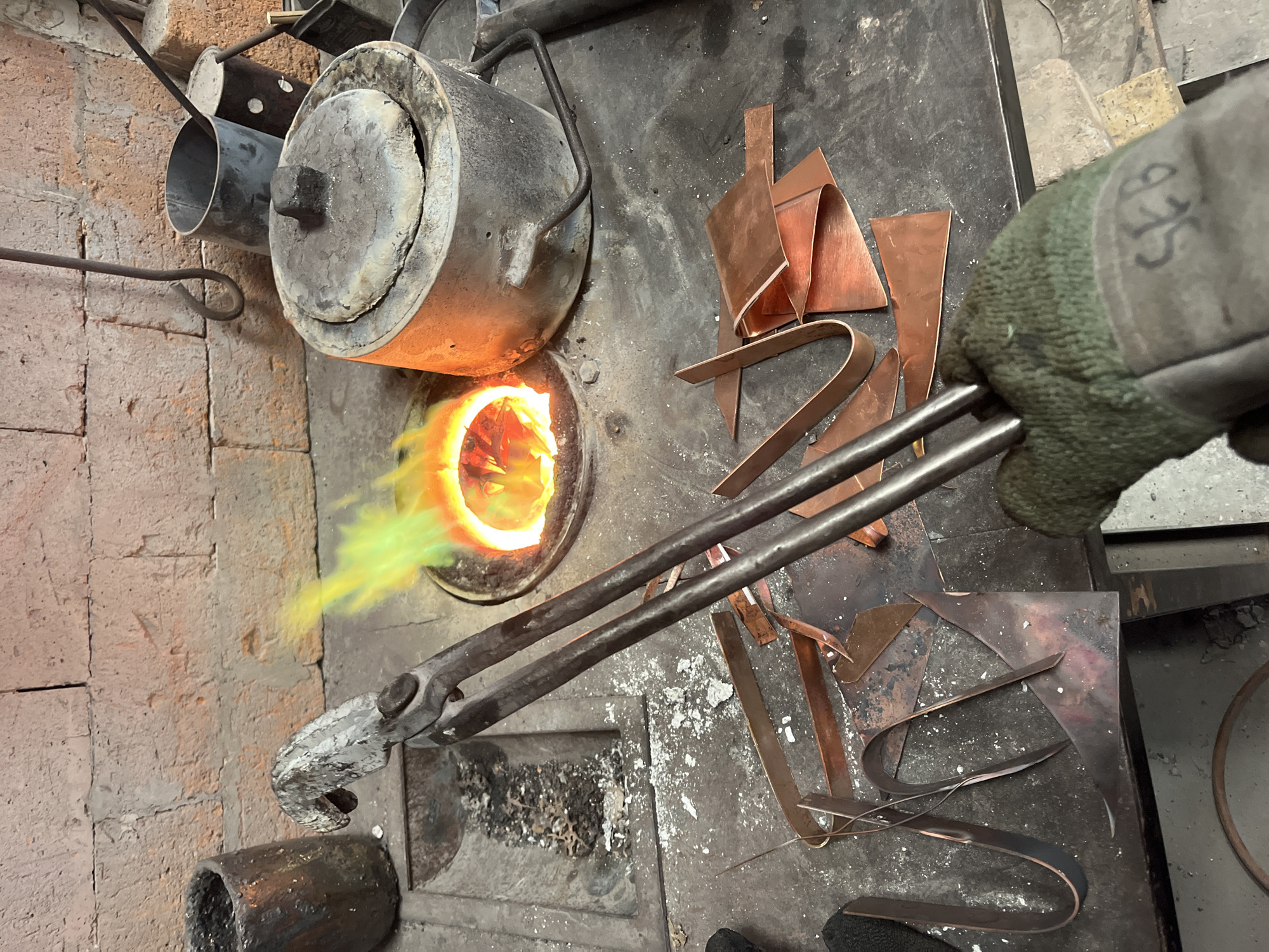 An overhead shot captures an industrial metalworking scene in Saint-Rémy-lès-Chevreuse, France. The central focus is a person's gloved hand, clad in a green and black work glove, holding a pair of long, sturdy metal tongs. The number "925" is visibly written in black marker on the back of the glove.
To the left, a brick structure houses a cylindrical metal furnace or forge, from which bright, vivid orange and green flames erupt, indicating intense heat. Adjacent to the furnace are various pieces of copper-colored metal, some appearing as flat sheets or strips, others bent into irregular shapes, scattered across a dark, grimy workbench or surface.
Further left, partially visible metal pipes or cylinders are integrated into the brickwork. The overall environment appears to be a workshop, characterized by the robust, well-used equipment and the scattered metal debris. The activity depicted suggests a process involving high-temperature metal treatment, likely melting or heating metal for shaping or casting, with the tongs poised for manipulation. The scene conveys a sense of focused, hands-on craftsmanship in a specialized environment.