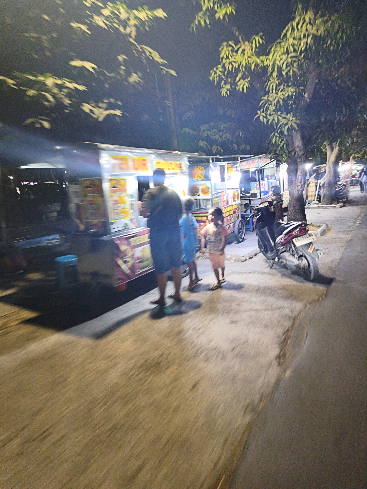 The image depicts a nighttime scene in Surakarta, Indonesia, focusing on a street-side food stall area. A prominent food cart, illuminated by bright lights, displays colorful food items. To the right of the cart, a person is partially visible, facing away from the camera. In front of the food cart, three children are present: one in a light blue shirt and shorts, another in a peach-colored dress, and a third, slightly further back, in a darker blue shirt. A dark-colored motorcycle is parked near the food cart. The environment suggests a public walkway or street with trees lining the background. The lighting is dim, with artificial light from the food cart and possibly streetlights. The image is blurry, indicating motion during the capture.