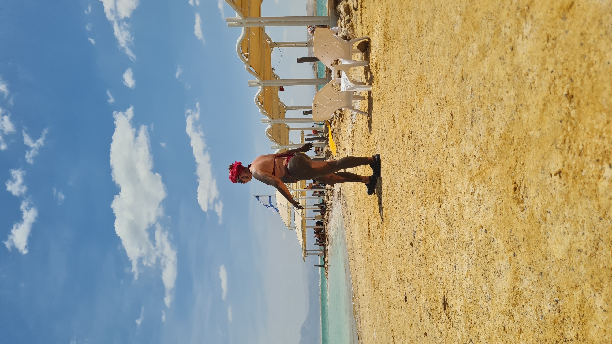 A man in a bathing suit and a red head covering is covered in mud and is holding a push-up position on a sandy beach. In the background are beach umbrellas and beach chairs. Behind that is a body of water and mountains in the distance. The sky is blue and partially cloudy. This scene is in Tamar Regional Council, Israel.