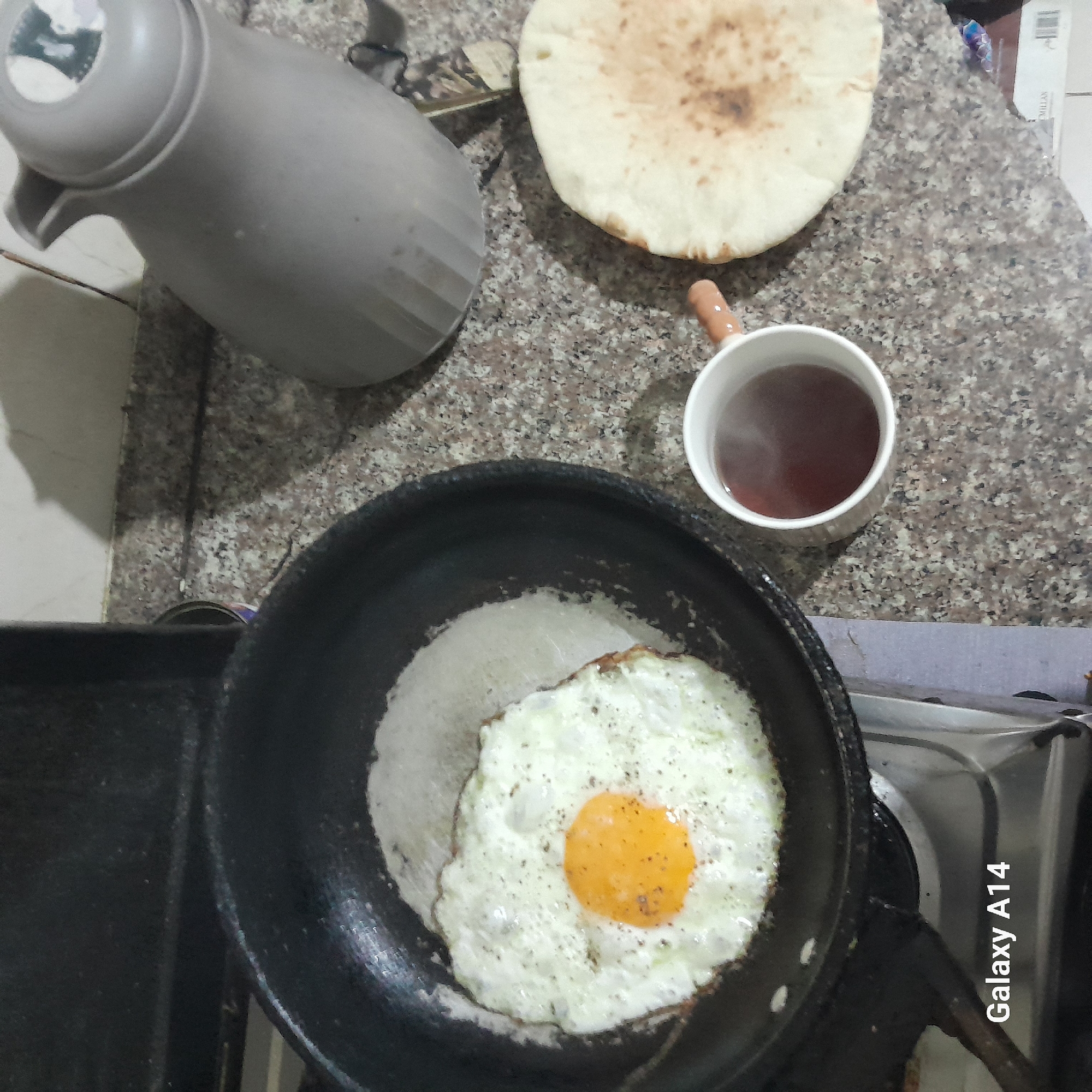 A close-up, high-angle shot presents a simple breakfast scene. The location is unknown, but appears to be indoors on a countertop or cooking surface. In the foreground, a black frying pan contains a fried egg. Adjacent to the pan, a silver gas stove with the text "Galaxy A14" printed on it is visible. In the background, there is a grey thermos, a piece of pita bread, and a white ceramic mug filled with a dark liquid, presumably tea or coffee. The countertop features a speckled grey and beige pattern. No people are directly visible in the frame. The scene suggests a quiet, solitary morning meal.