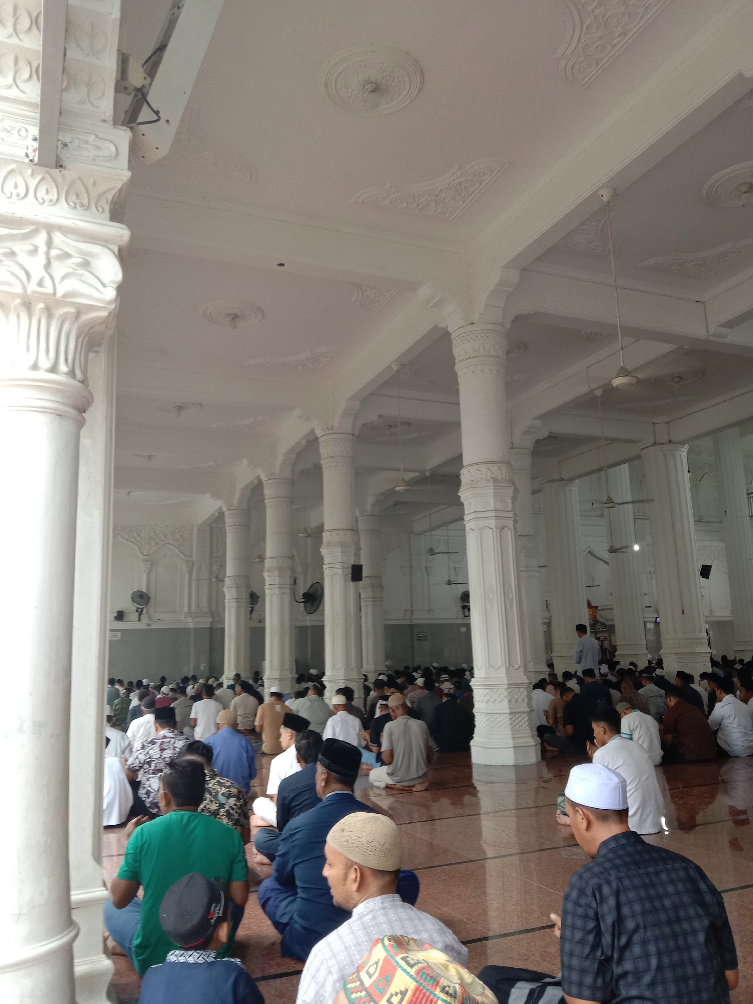 This image depicts the interior of a mosque, likely in Lhokseumawe, Indonesia, during a prayer gathering. Numerous men, dressed in traditional attire and skullcaps, are seated on a polished floor, facing towards a central point that is not fully visible. The mosque features ornate white columns and a decorative ceiling with intricate patterns. Ceiling fans are visible, indicating a warm climate. The overall atmosphere appears to be one of communal worship and serenity.