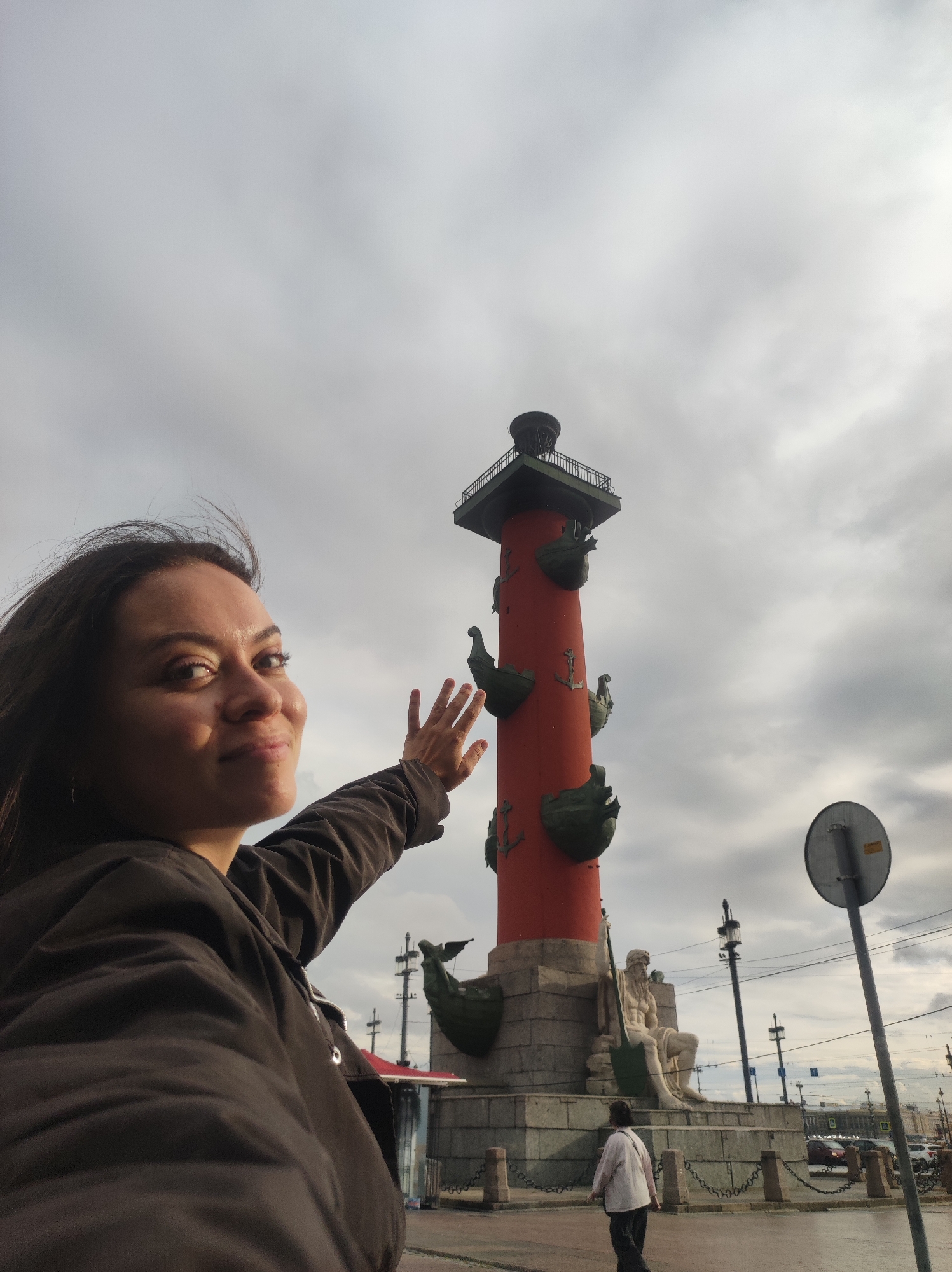 In Saint Petersburg, Russia, a woman with dark hair, wearing a brown jacket, poses for a selfie in front of the Rostral Columns on Vasilyevsky Island. She is smiling and extending her left hand towards the column, which is tall, red, and adorned with green nautical figures and anchors. Behind her, the cloudy sky is visible, along with the base of the column featuring stone sculptures. A street sign and lampposts are also present in the background, along with a pedestrian walking away from the camera.