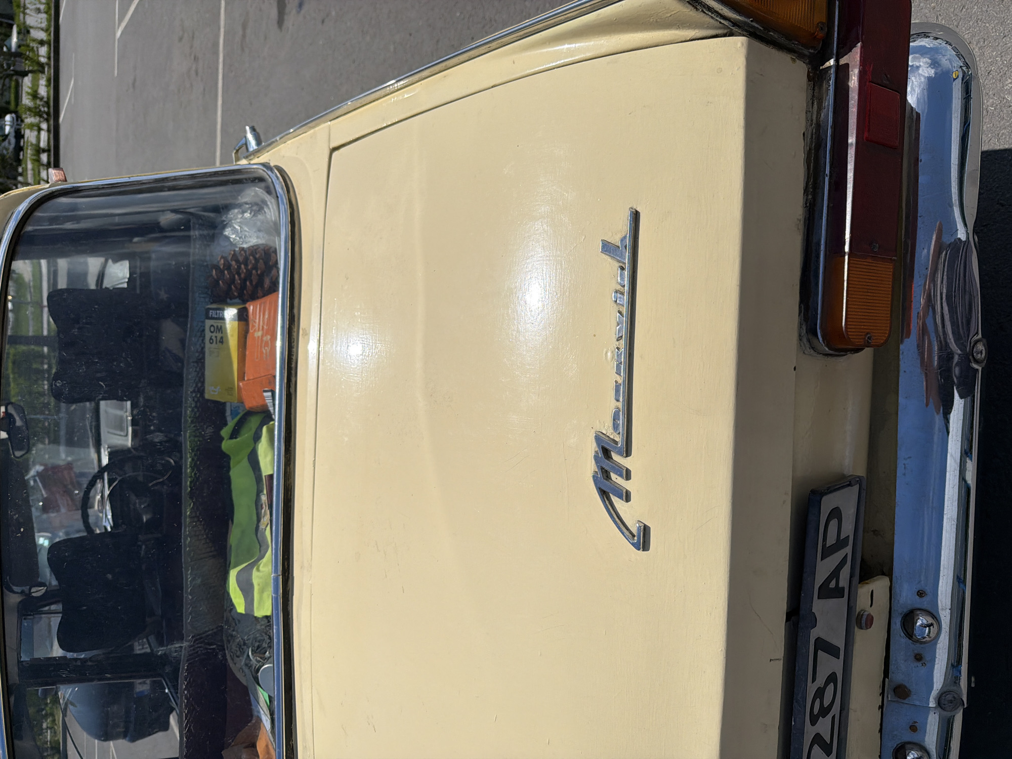 The image shows the rear right portion of a light yellow "Moskvich" car. The car is parked on asphalt, possibly in a parking lot. The "Moskvich" emblem is visible on the rear panel. A Bulgarian license plate "287 AP" is attached to the rear, and a chrome bumper with lights is visible. Reflected objects in the car's interior, including items like a pine cone, filter box, and reflective vest, indicate the car's interior is somewhat cluttered.