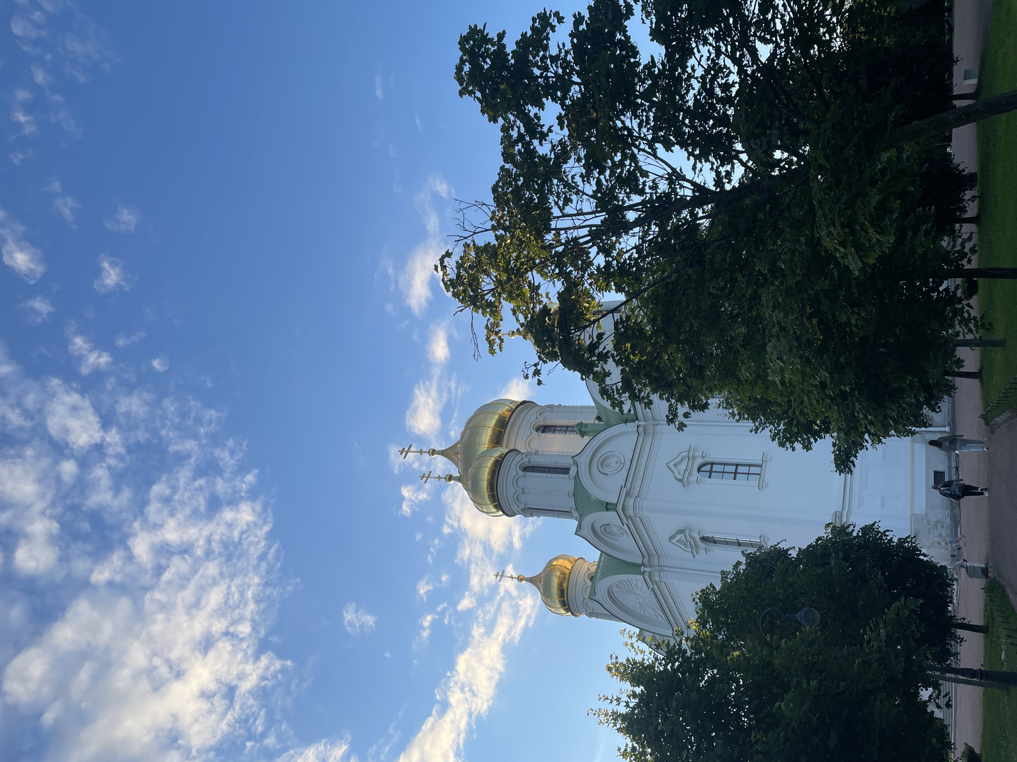 This is a full-frame, low-angle shot of the Alexander Nevsky Cathedral in Pushkin, Russia, under a partially cloudy blue sky. The white, neoclassical cathedral with green trim and multiple golden domes dominates the upper left portion of the frame. Three gold domes with crosses are visible. Lush green trees, casting shadows, frame the right and lower edges of the image, with some foliage obscuring parts of the cathedral's base. In the lower portion of the image, a paved pathway leads into the scene, with two people walking away from the camera towards the cathedral. A lamppost is visible on the left side of the pathway, and a trash receptacle is situated near the pathway on the right. The ground is covered in grass.