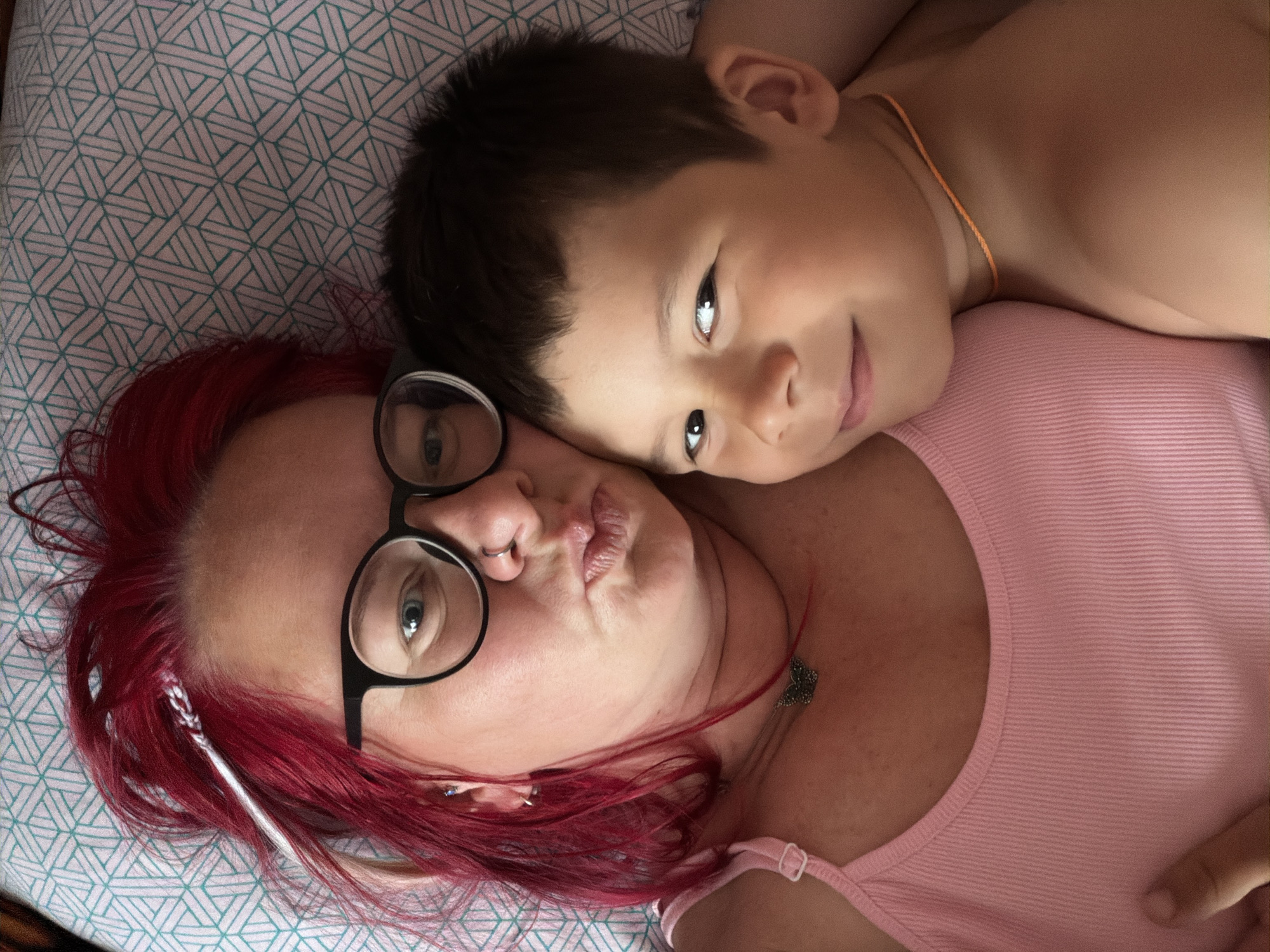This is a close-up, overhead shot of a woman and a young boy lying down together, possibly on a bed given the patterned fabric visible beneath them. The woman has bright red hair and is wearing black-rimmed glasses. She has a piercing on her nose and a necklace with a butterfly pendant. The boy, who appears to be younger, has dark hair and is resting his head on the woman's shoulder. He is wearing a thin orange cord necklace. Both individuals are looking towards the camera. The boy has a gentle smile, and the woman's expression is more neutral, with her lips slightly pursed. The overall tone of the image is intimate and affectionate. The lighting suggests it is daytime. There is no visible text in the image.