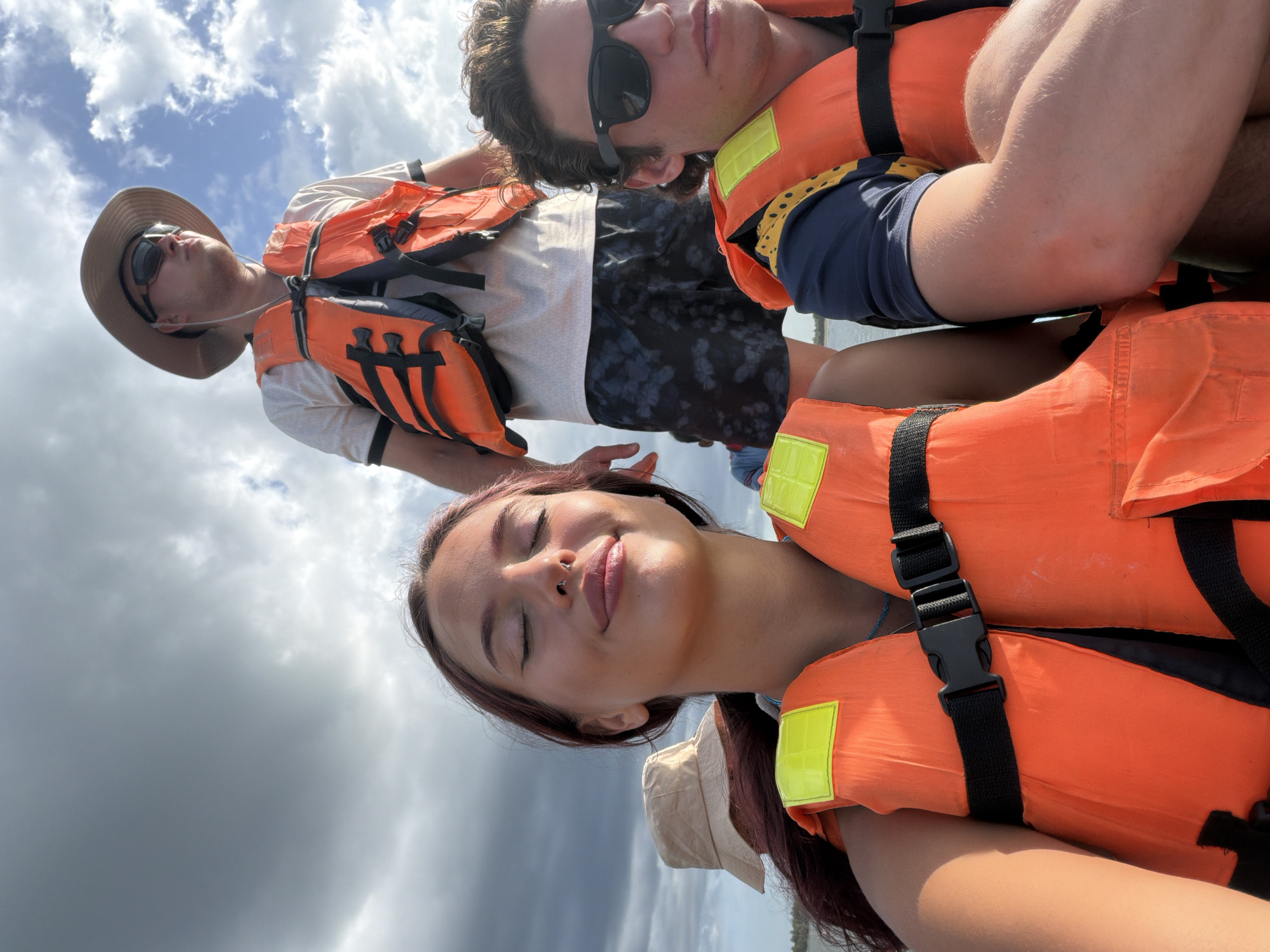 In Pottuvil, Sri Lanka, a group of four individuals, two women and two men, are on a boat under a cloudy sky. The woman in the foreground, with her eyes closed and a relaxed smile, wears an orange life vest. To her right, a man with sunglasses and a dark t-shirt also wears an orange life vest. Behind them, a man in a hat and sunglasses stands, also in an orange life vest. Another person wearing a bucket hat is partially visible to the left of the foreground woman. The group appears to be enjoying a boat ride, with the dark, dramatic clouds suggesting potential weather changes.