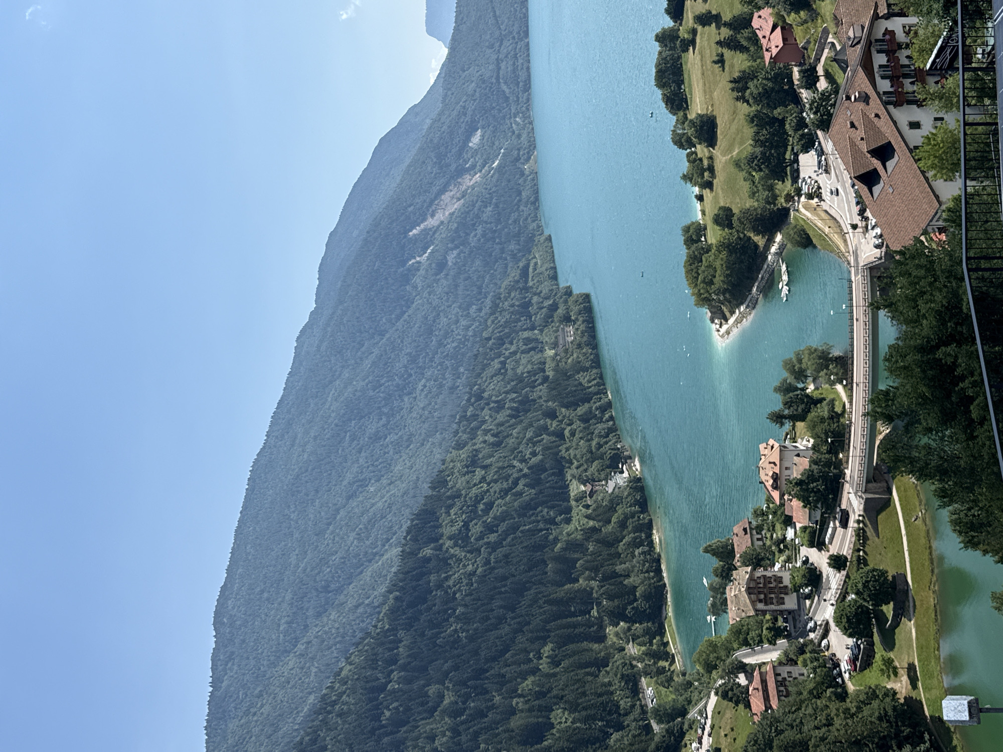 The image captures a panoramic view of a serene lake and a charming Italian village nestled at its edge, likely in Molveno, Italy. The expansive, turquoise-blue lake dominates the foreground and midground, its waters shimmering under a clear, bright sky. A lush, green mountain range rises majestically behind the lake, its slopes covered in dense forestation.
The village, situated on the lakeshore, features a cluster of buildings with characteristic terracotta roofs. A bridge spans a section of the lake, connecting different parts of the settlement. Cars are visible on the roads and in parking areas, suggesting a peaceful, populated scene. Boats are scattered on the water, some docked near the shore and others out on the lake. The overall impression is one of tranquility and natural beauty, with the vibrant colors of the water, greenery, and architecture creating a picturesque landscape.