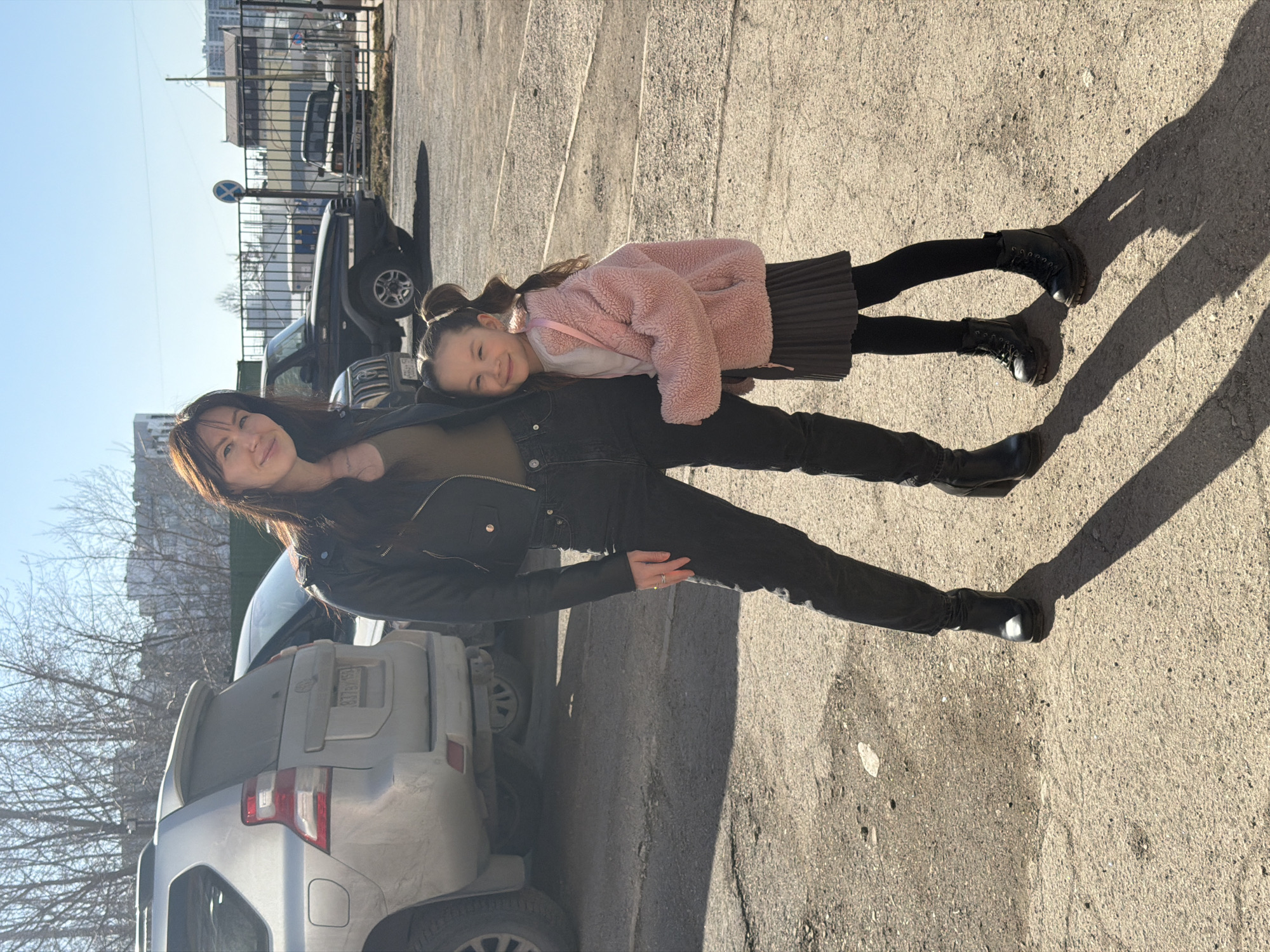 A young girl in a pink coat lies on the back of a woman in a black jacket and jeans. They are standing on a concrete surface in what appears to be a parking area. Several vehicles are visible in the background, including a silver SUV parked directly behind the woman. The location is in Perm, Russia. The time of day is daytime.