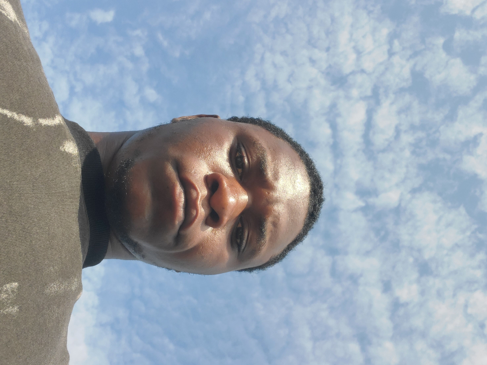 A low-angle, close-up portrait, presumably taken in Winters, United States of America, depicts a young adult male. He has dark skin, short, tightly curled black hair, and a light amount of facial hair on his jawline and chin. His face shows a sheen, consistent with sweat or direct illumination, particularly on the forehead, nose, and cheeks. He maintains a direct gaze towards the camera with a neutral, unsmiling expression. He is dressed in a dark-colored, likely black or dark grey, short-sleeved crew-neck shirt that features faint, irregular lighter streaks or patterns on the chest and shoulder. The background consists of a clear blue sky with scattered white cumulus clouds, indicating bright daytime conditions.