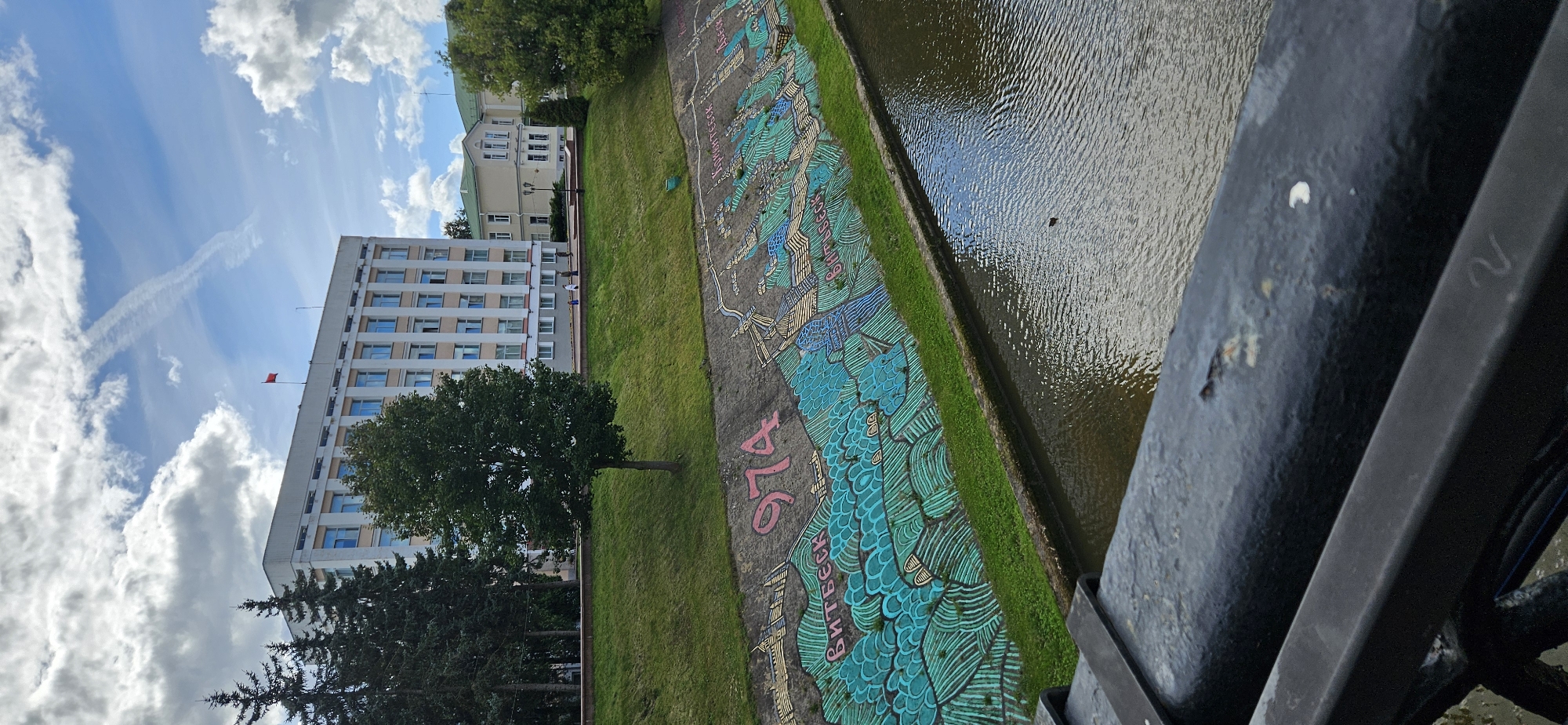 This image captures a scene in Vitebsk, Belarus, featuring a wide, calm body of water in the foreground, likely a river or canal, with a dark metal railing partially obscuring the view. Along the bank opposite, a concrete retaining wall is adorned with a vibrant mural. The mural depicts a stylized landscape with blue and green wavy patterns representing water and land, overlaid with images of buildings and bridges. The number "974" is prominently displayed in pink lettering on the wall, above the mural. Further back, a grassy embankment slopes upwards, leading to a row of trees and several buildings. The most prominent building is a large, light-colored structure with multiple floors and numerous windows, topped by a red flag. Other, smaller buildings with similar architectural styles are also visible. The sky above is partly cloudy with a contrail from an aircraft. A few people are walking on a paved path behind the embankment, in front of the buildings. The overall impression is an urban landscape where art and architecture are integrated with a natural water element.