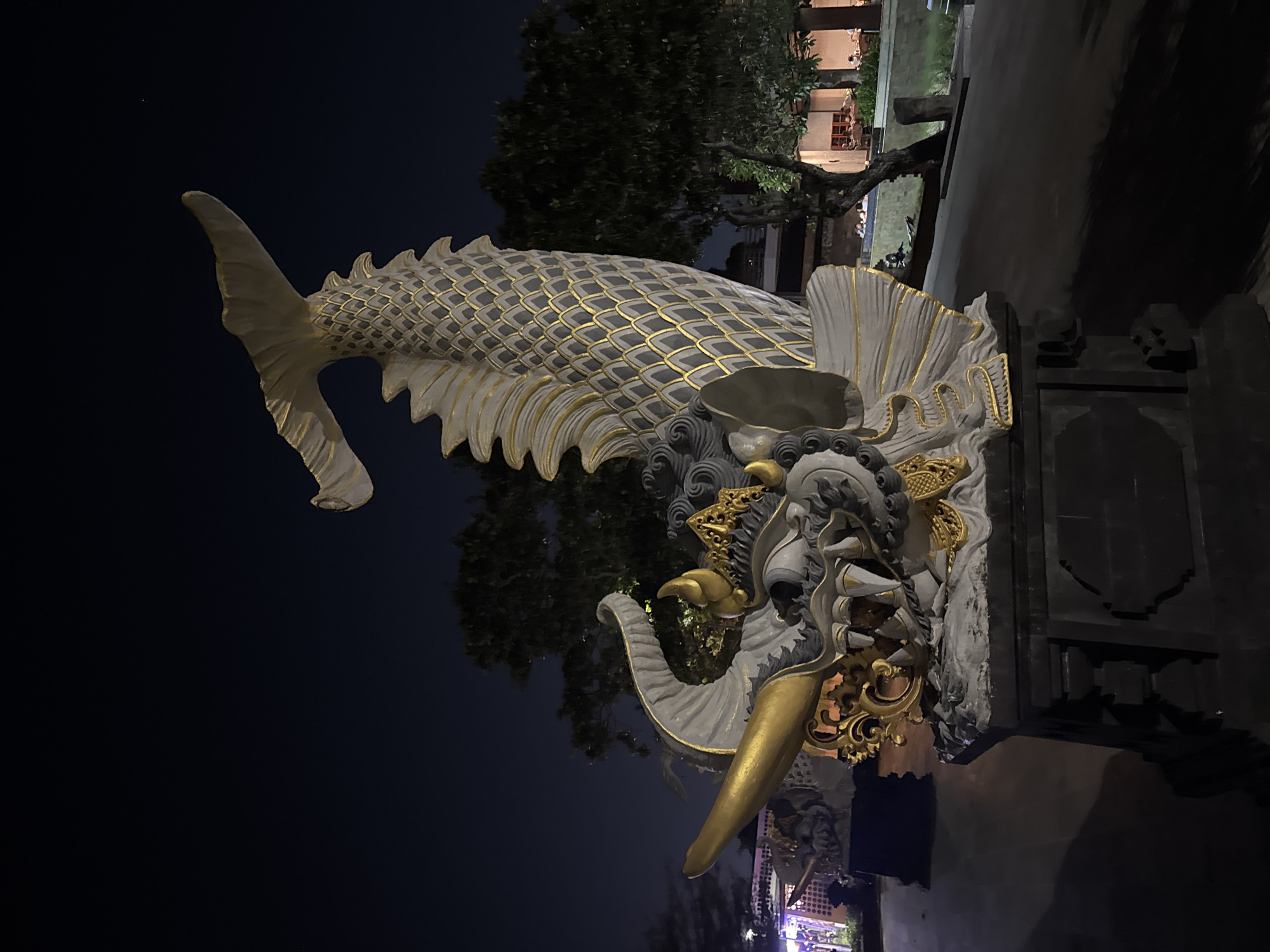 A statue of a mythical creature, possibly a dragon or sea serpent, is depicted at night. The creature is white with gold accents, featuring intricate scales, sharp teeth, and a long, curved horn. It is mounted on a dark, stone pedestal. The statue is located in an outdoor area, with trees and buildings visible in the background. The sky is dark, indicating it is nighttime. The overall impression is one of grandeur and artistry. Based on the architectural style and the presence of trees typical of the region, this statue is likely located in Kuta, Indonesia, a popular tourist destination known for its vibrant culture and temples.