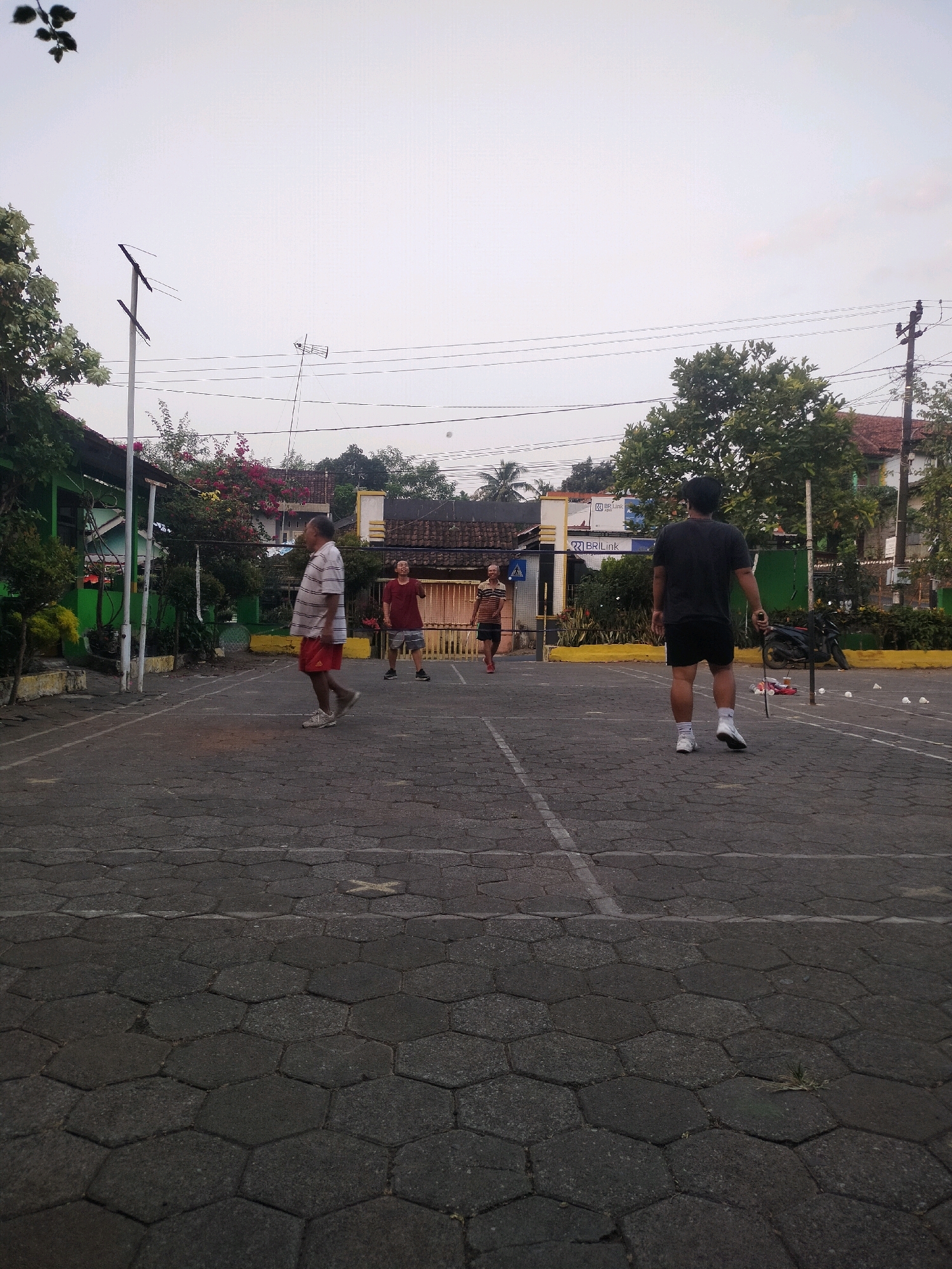 The image depicts an outdoor badminton court in Boja, Indonesia, likely during late afternoon given the soft lighting and the shadows. The court surface is paved with interlocking hexagonal stones. In the foreground, the court lines are visible. There are four men on the court, engaged in a badminton game.
On the left side of the court, an older man in a striped t-shirt and red shorts is walking. Further back on the left, a man in a red t-shirt and grey shorts is playing. To his right, another man in a red t-shirt and darker shorts is also playing. On the right side of the court, facing away from the camera, is a man in a black t-shirt and black shorts, holding a badminton racket.
In the background, there are buildings, trees, and power lines. One building has a green facade and a yellow border. Another building in the distance has a red tile roof. There are also several shuttlecocks visible on the court, some scattered near the man with the racket on the right, and a few more further down the court. A motorcycle is parked on the right side of the image, near the edge of the court. The sky is overcast and bright.
