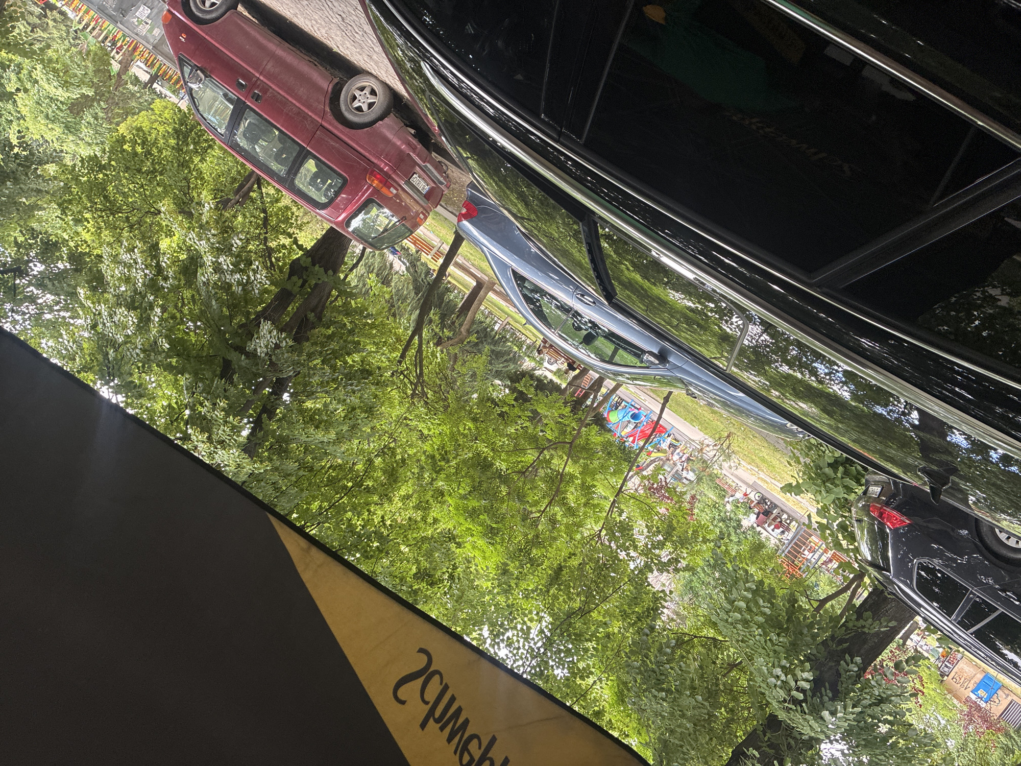 Here is a description of the image:
The image appears to be taken from an outdoor location, possibly beneath an awning or umbrella, as indicated by the dark and light yellow material in the lower-left portion of the frame. The main subject is a parking area with several vehicles. A maroon van is parked on the left side of the frame, its license plate visible, and a blue sedan is visible through the reflection on a glossy black car. In the background, there is a park with trees and a colorful children's play structure. There are benches, suggesting a recreational area. The lighting indicates it is likely daytime. The city is Unknown, Unknown.
