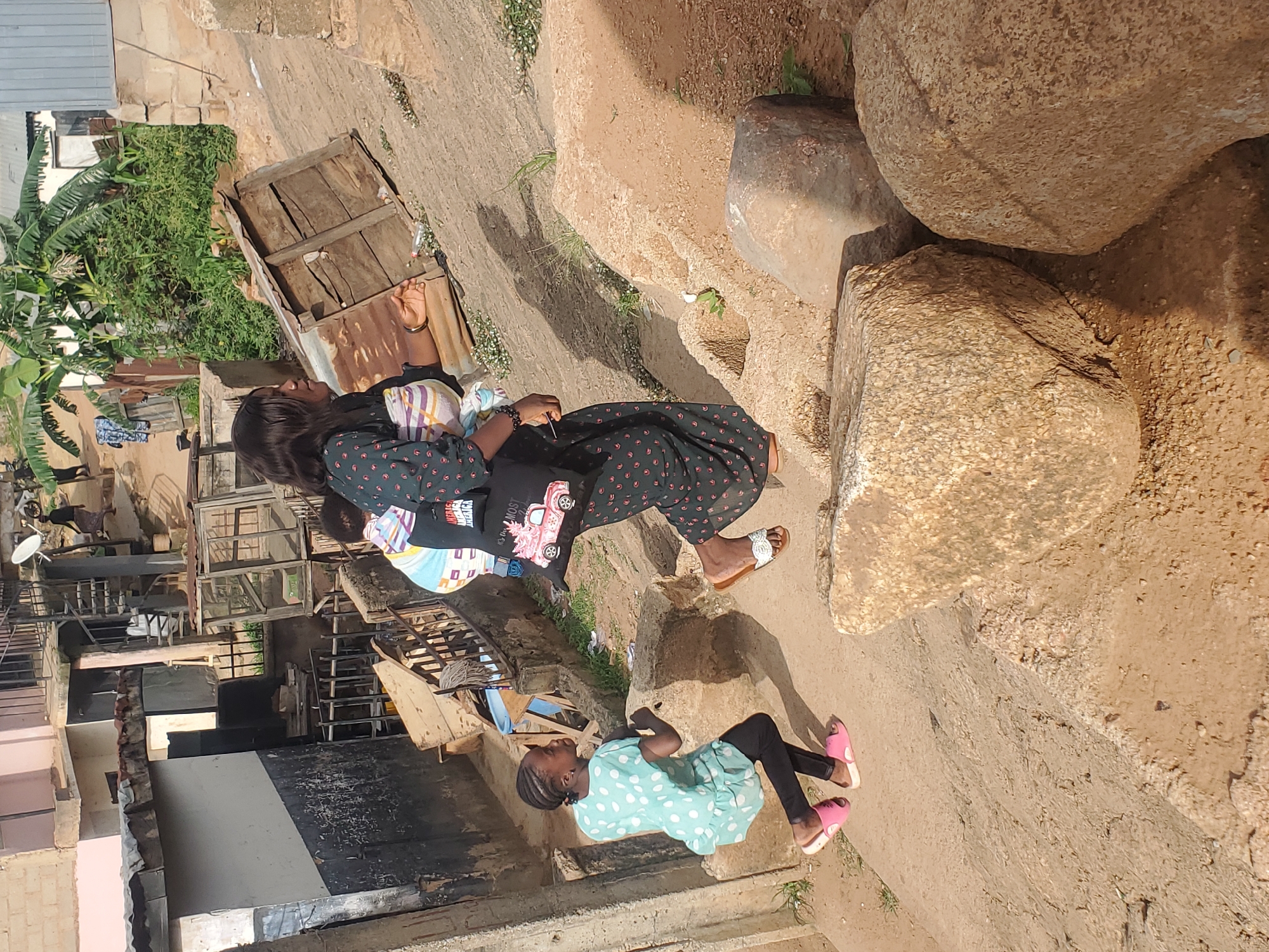 In Gwong, Nigeria, a young girl with braided hair and a light blue polka-dot dress runs up a dirt path. She wears pink slip-on sandals and black leggings. Behind her, a woman in a dark patterned dress carries a baby on her back. The baby is wrapped in a multi-colored blanket. The woman also carries a black tote bag with a pink cartoon car graphic. The background shows a hilly, unpaved environment with several rustic buildings and vegetation. The scene is illuminated by natural daylight, casting shadows on the ground.