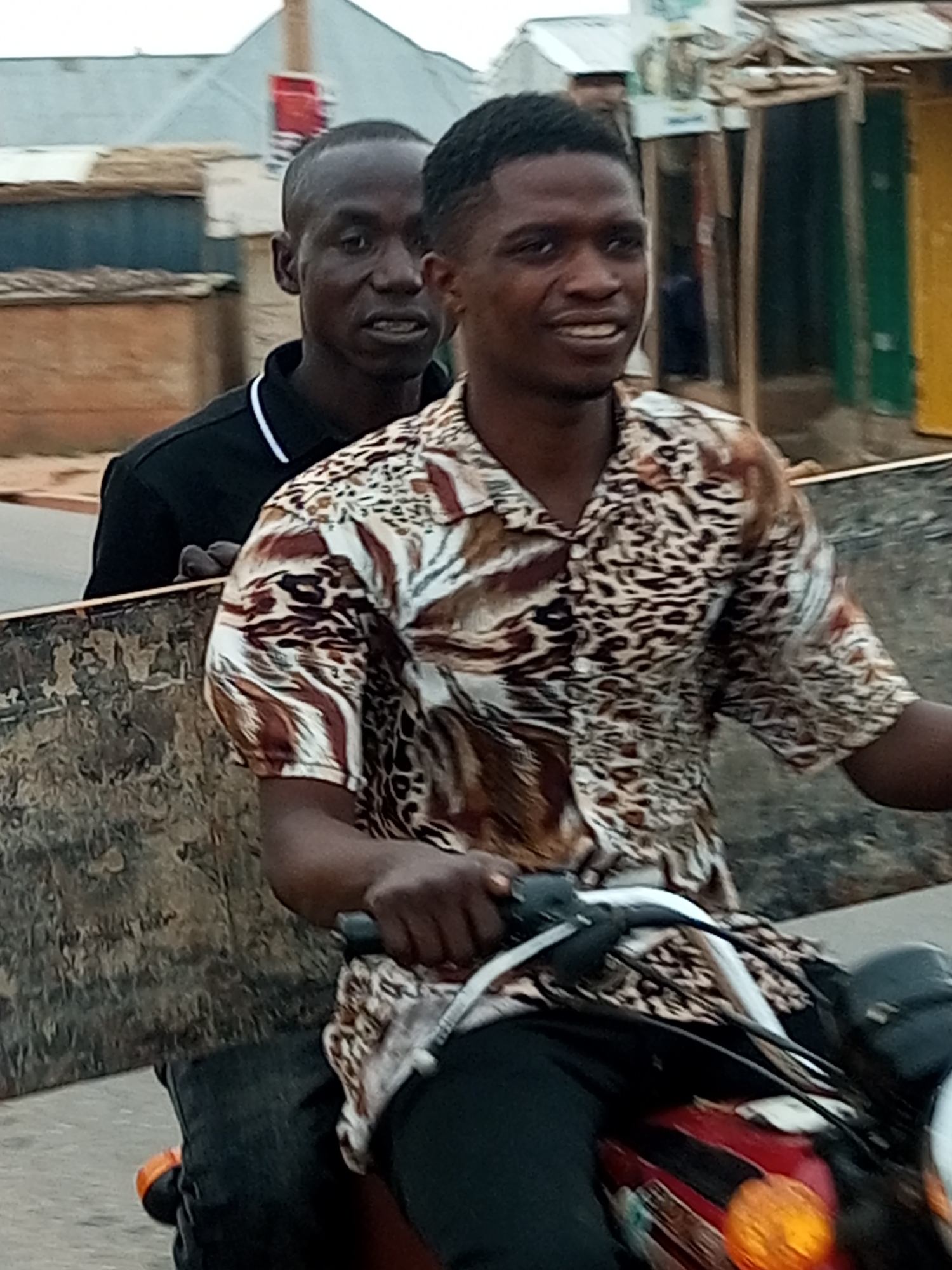 Two men are on a motorcycle, with the driver in the foreground wearing a leopard-print shirt and dark pants, and the passenger behind him in a black shirt. The motorcycle has a red fuel tank and orange and white accents. The scene appears to be in a roadside area with buildings and structures visible in the background. The overall setting suggests a street in the city of Gombe, Nigeria, likely in a commercial or residential area.