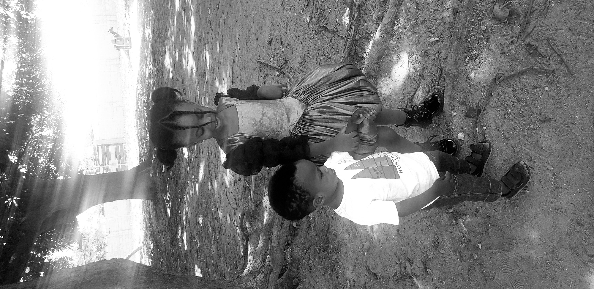 This black and white photograph, taken in Narayi, Nigeria, depicts two children standing on a dirt ground under dappled sunlight. On the right, an older child, appearing to be a girl with dark skin and braided hair styled into two buns with hanging braids, wears a dress with a light-colored, possibly metallic, bodice, dark puffy sleeves, and a voluminous skirt. She wears dark, shiny combat-style boots. Her right hand grasps the left hand of the younger child. The younger child, positioned on the left, appears to be a boy with dark skin and short dark hair. He wears a light-colored t-shirt with a graphic design featuring text "SK-470" and "NORTH," dark jeans, and dark sandals. Both children are looking downwards. The ground is composed of uneven dirt with visible roots and scattered debris. Large tree trunks and exposed roots are present on the left and in the background. Bright sun rays filter through the tree canopy from the upper right, casting strong highlights and shadows. In the overexposed background on the right, the faint outline of a structure, possibly a wall or fence, is discernible, along with a large animal, potentially a camel or cow, lying down.