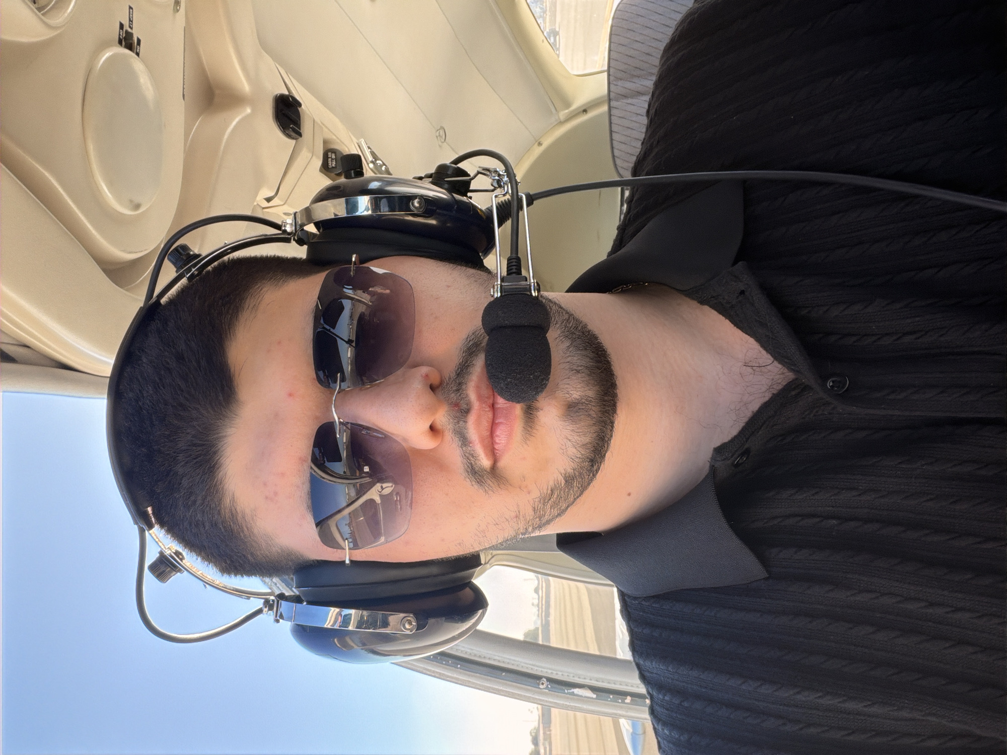 A man wearing a black collared shirt and aviator sunglasses is inside an aircraft. He has headphones with a microphone on. The interior of the aircraft is visible, with beige panels and a striped seat in the background. Outside the aircraft window, a runway and clear blue sky are visible, suggesting an airport setting. The lighting indicates it is daytime.
