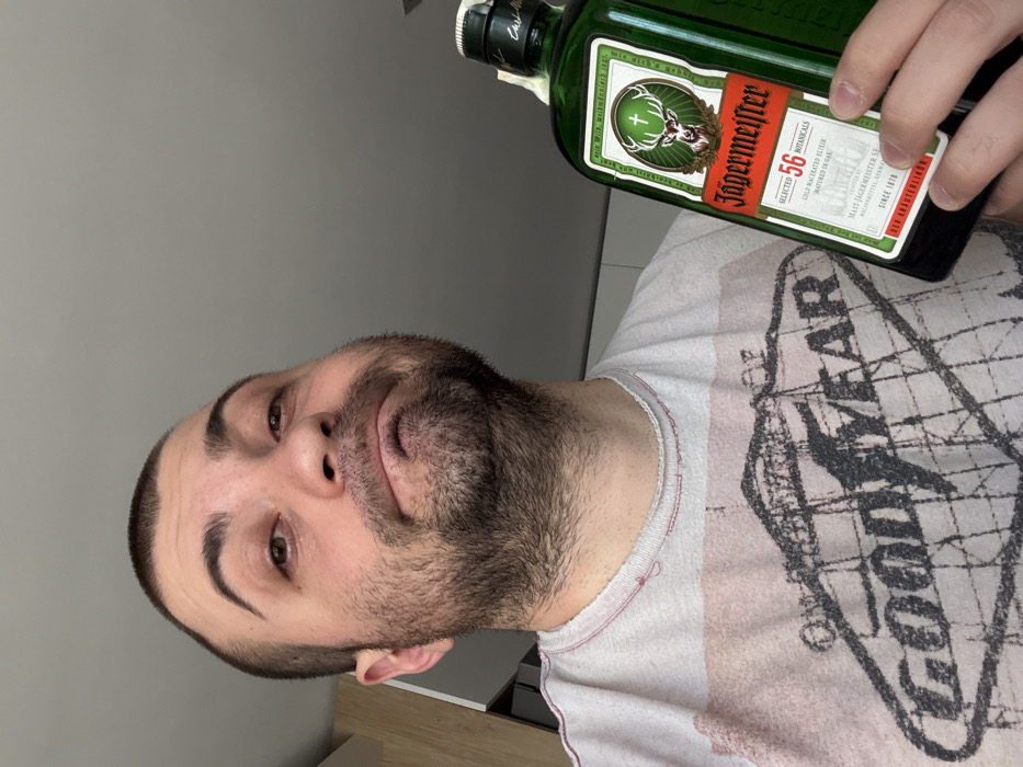 An adult male, with short dark hair and stubble, is depicted from a low angle, looking upwards towards the camera with a slight frown or pout. He wears a light-colored t-shirt with a faded black 'GOOD YEAR' graphic and a red-stitched, distressed collar. His right hand holds a green glass bottle of Jägermeister, prominently displaying its label. The background is a soft-focus indoor environment, featuring a light grey wall or ceiling and a light wooden or laminated cabinet to the left. The scene is located in Sofia, Bulgaria.