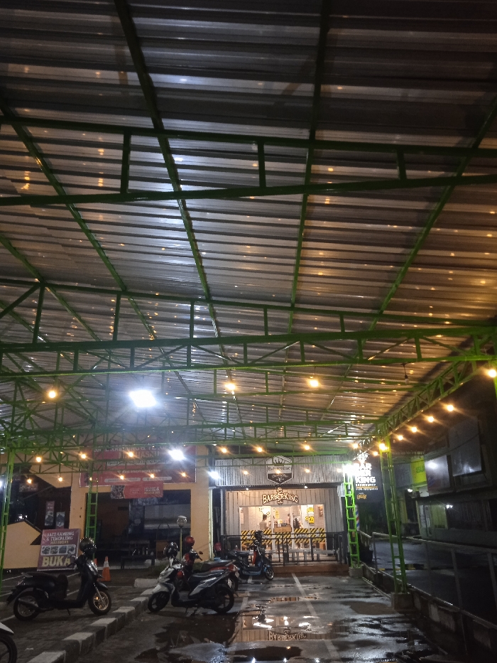 This nighttime image captures an outdoor commercial area, likely a food court or a strip of small businesses, located in Kecamatan Kartasura, Indonesia. The scene is dominated by a large, open-air canopy featuring a green metal framework supporting a corrugated metallic roof. Numerous warm string lights are strung across this structure, illuminating the space below.
The ground is paved and appears wet, possibly from recent rain, with white painted lines indicating parking spots. Several motorbikes of various makes are parked in the foreground, suggesting customer or staff presence.
On the left side of the frame, a food stall or restaurant is visible, marked by signs and banners. A prominent sign partially reads "Sate Kambing & Tengkleng" (Goat Satay & Tengkleng), and another sign below it clearly states "BUKA" (Open). A large red banner with white text is displayed above this establishment.
To the right, a modern barbershop named "BARBER KING" is clearly visible, with its distinctive logo and white-and-black striped facade. Large glass windows allow a view into its interior. The artificial lighting against the dark backdrop confirms the time of day as night. The overall atmosphere suggests a lively evening commercial setting.