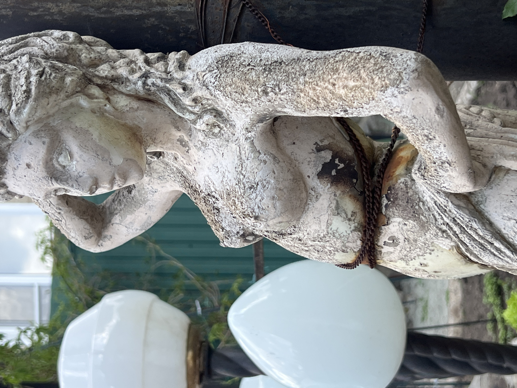 A weathered, light-colored statue of a nude female figure with flowing hair is displayed outdoors. A rusty chain is wrapped around the statue's midsection. Below the statue are several white globe-shaped objects that appear to be lamps. The background includes green vegetation, a metallic fence, and part of a white building. This image was taken near the city of Boguchar, Russia.